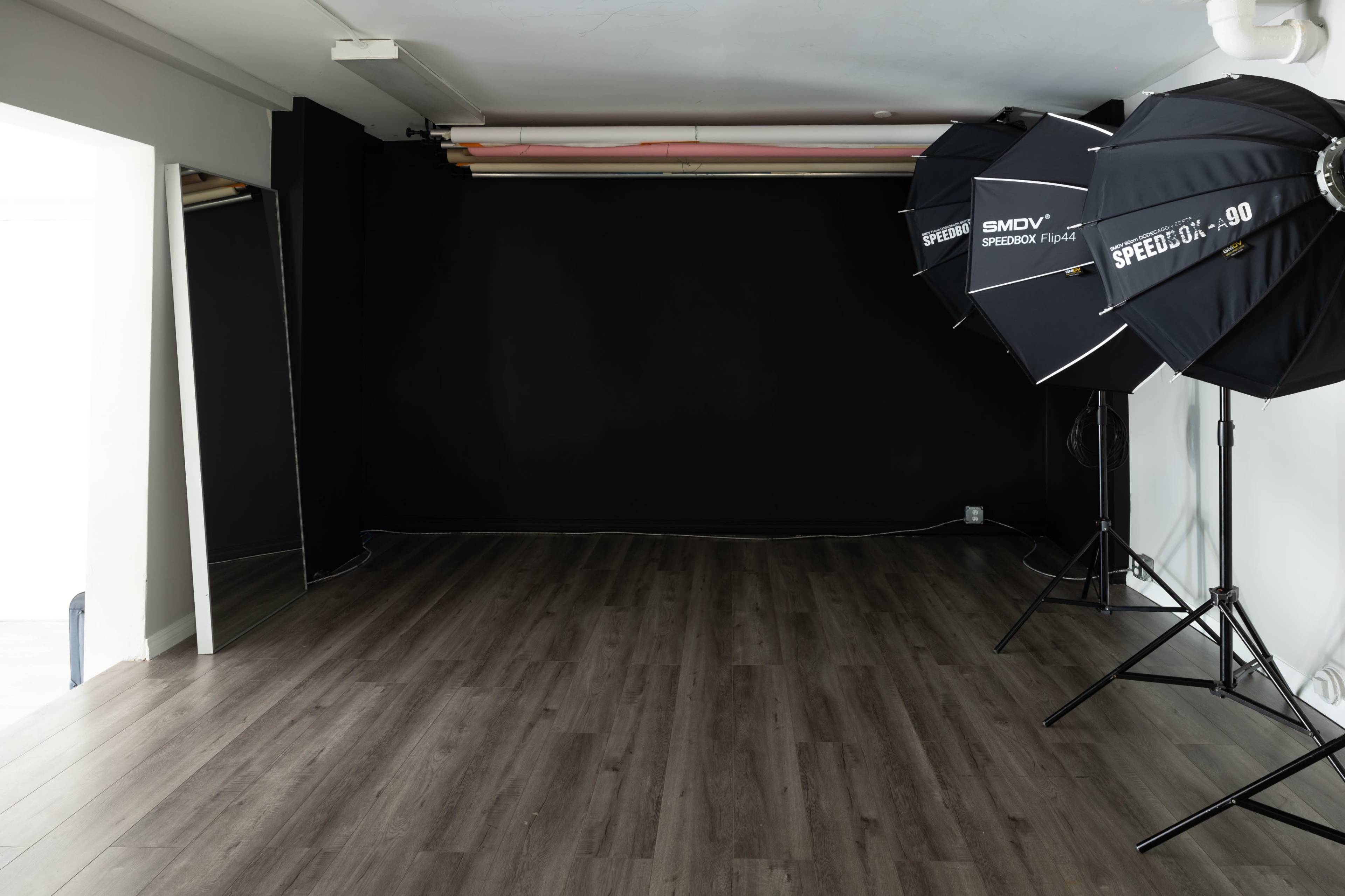 A spacious, empty studio room with wooden floors, a black wall, and two large photography softbox lights on tripods.