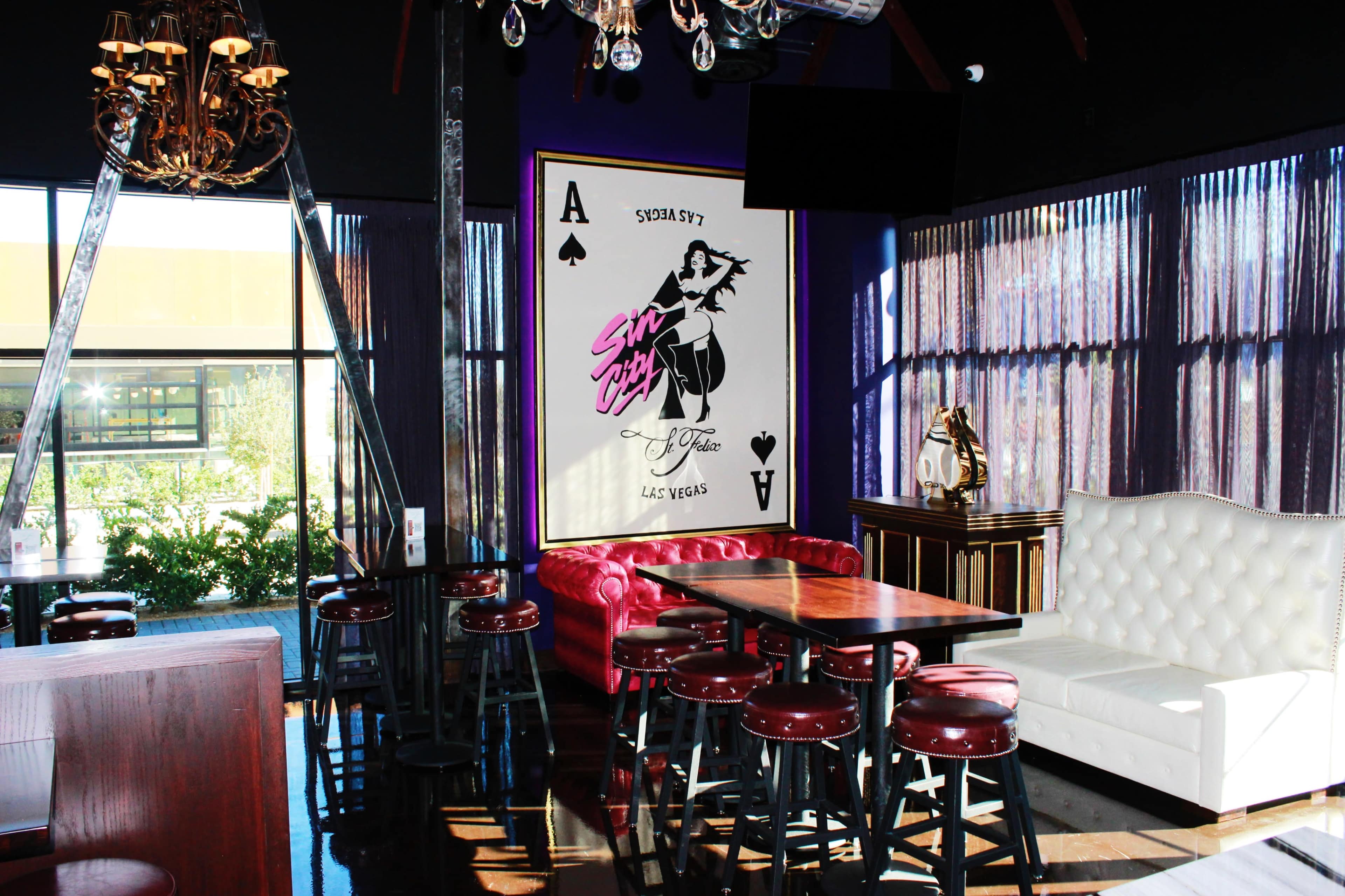 The image shows a stylish interior of a lounge featuring a red leather sofa, white seating area, tall tables, and a large playing card mural on the wall.