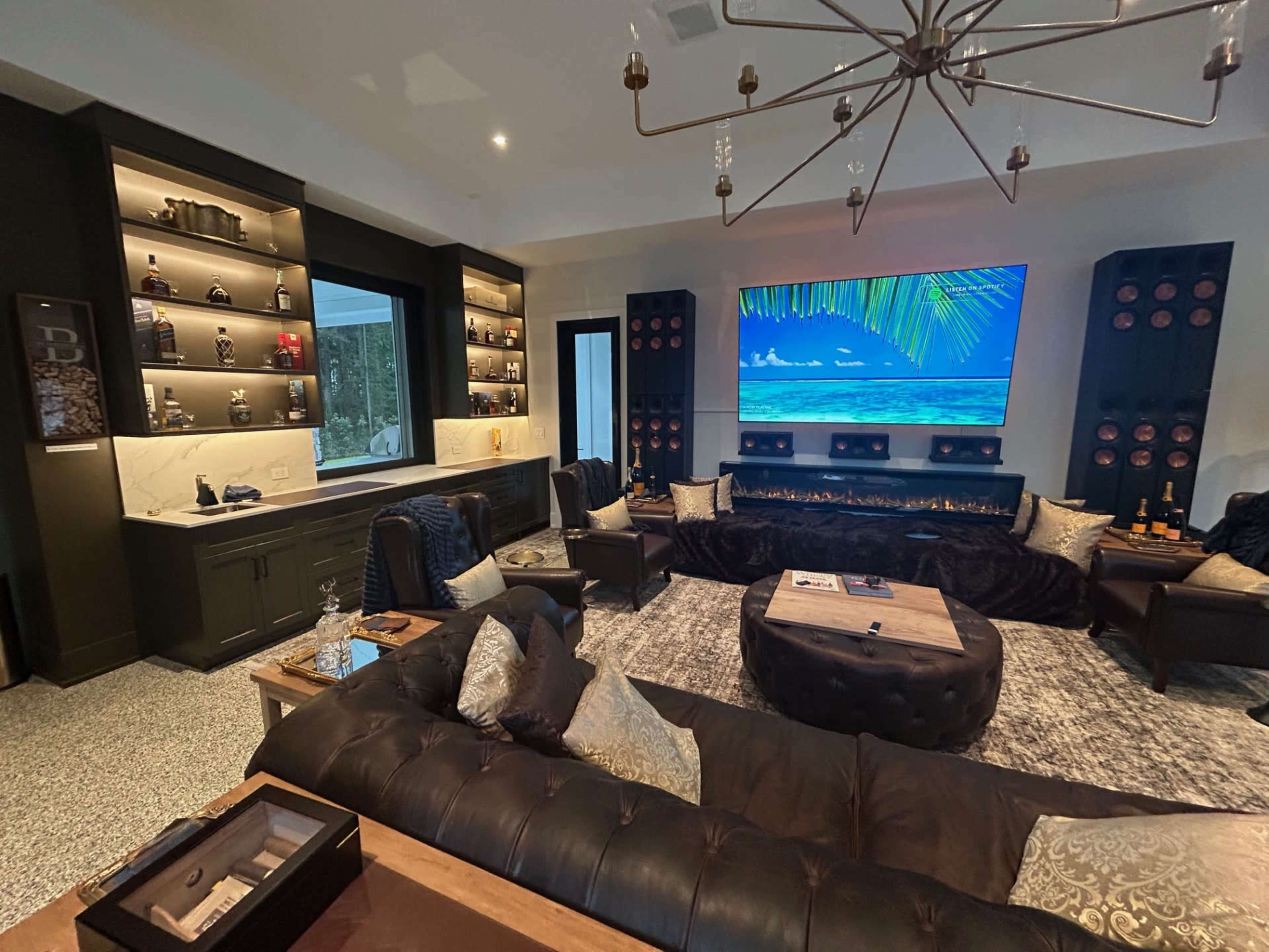 The image shows a modern living room with dark furniture, a large television mounted on the wall, and shelves displaying decorative items and wine bottles.
