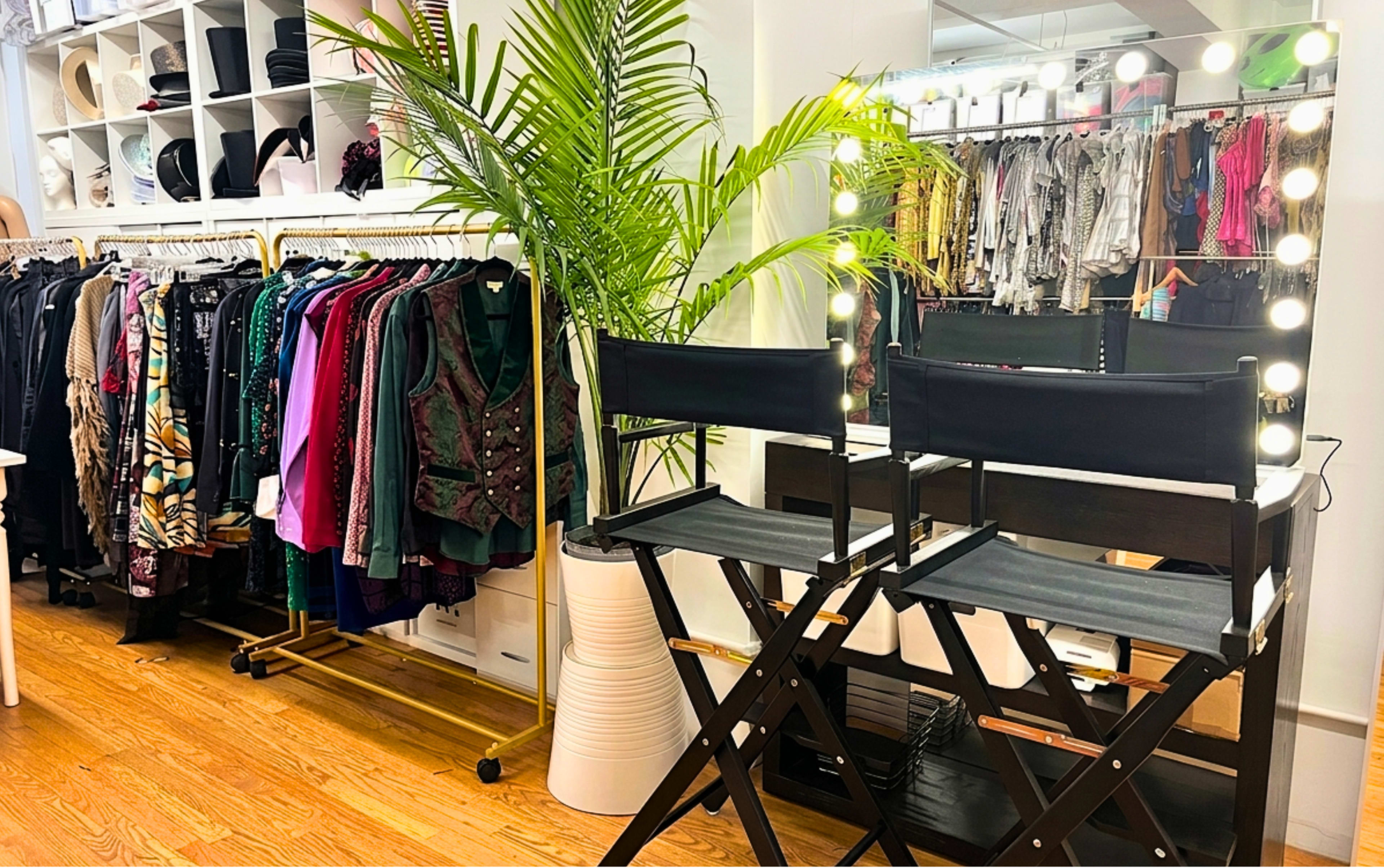 The image shows a modern dressing room setup with two black director's chairs facing a lighted mirror, surrounded by a rack of colorful clothing and a large potted plant.