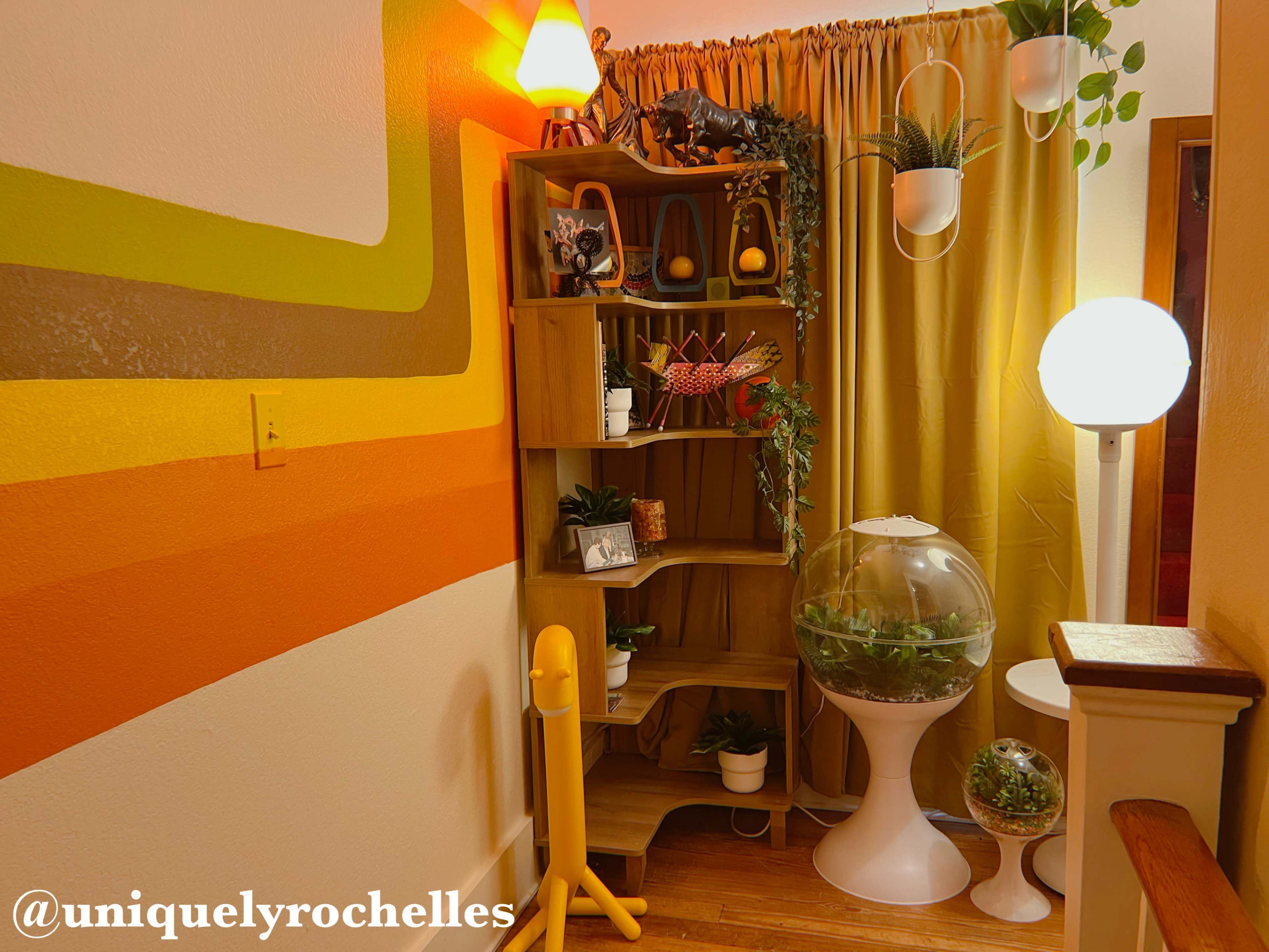 The image shows a cozy corner of a room featuring a wooden shelf with plants, a globe planter, a lamp, and colorful wall decor.