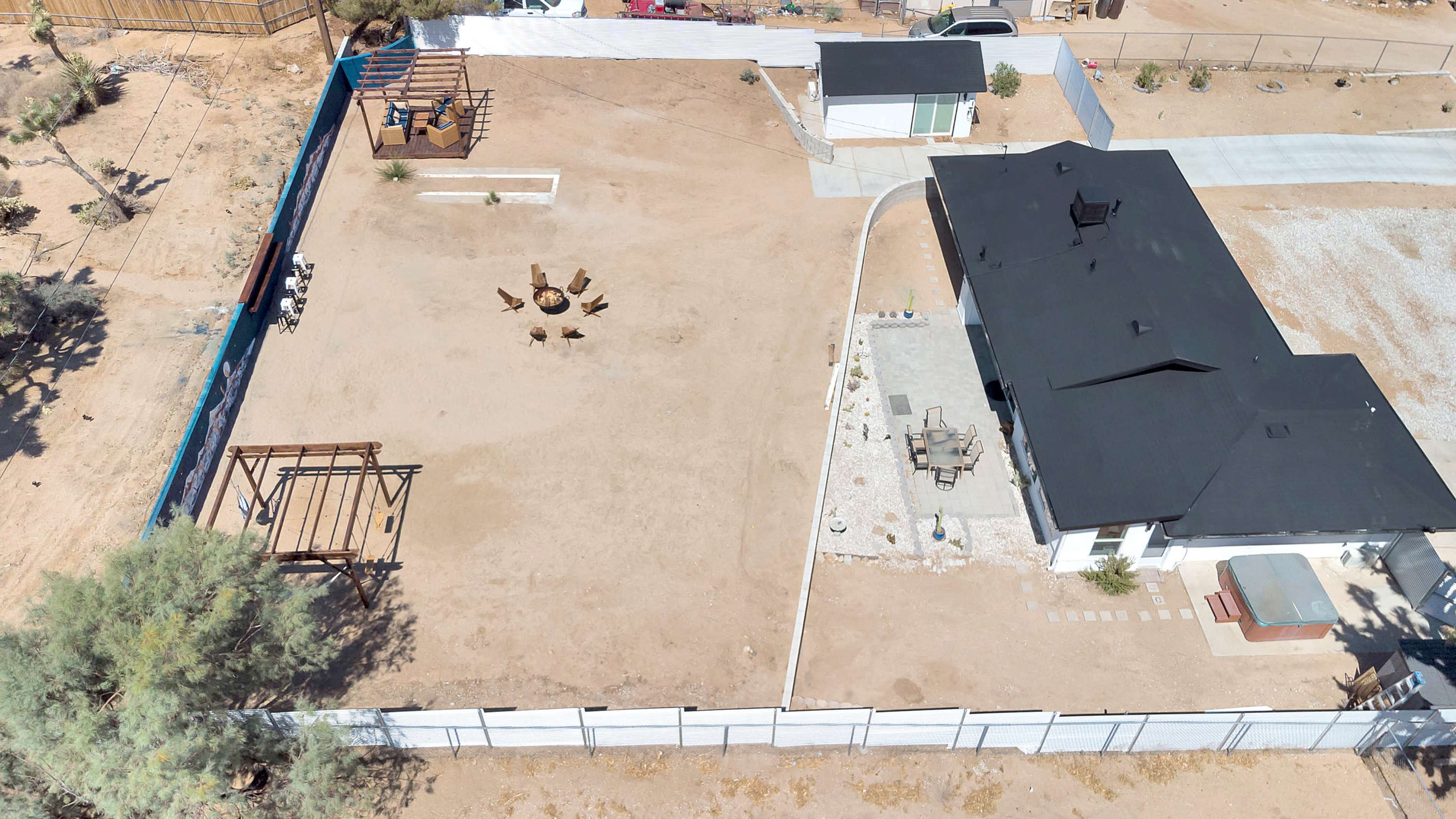A residential property with a large, empty yard featuring a fire pit in the center, surrounded by a fence, and a house with a black roof on one side.