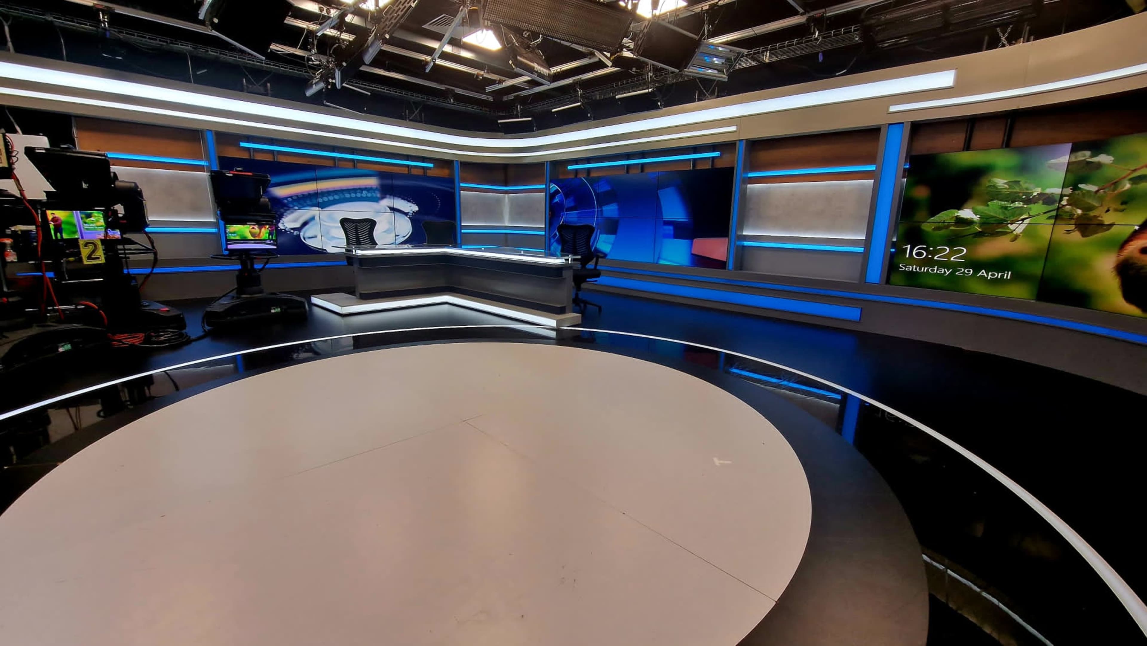 The image shows a modern television studio with a circular floor, multiple cameras, and screens displaying graphics and a digital clock.