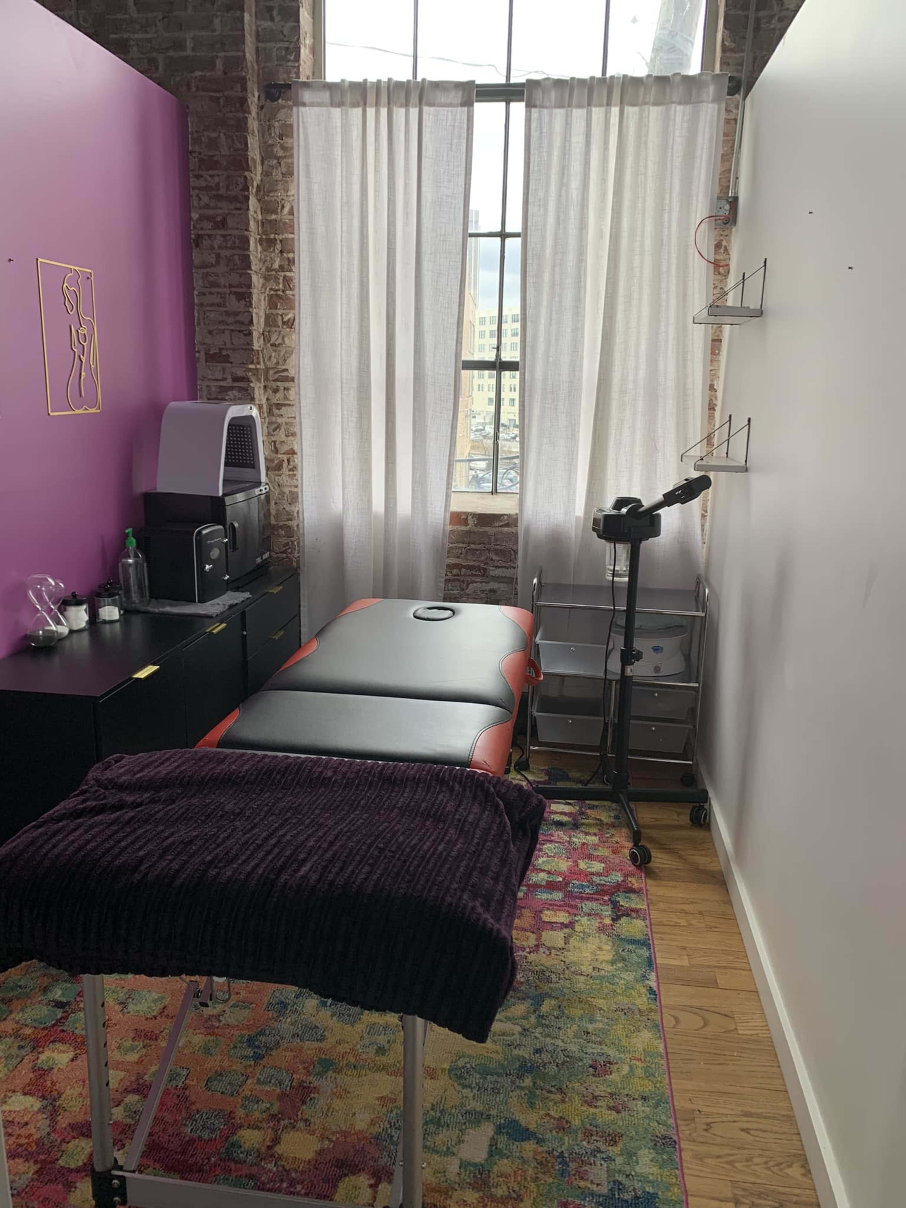A treatment room with a massage table, a small storage cart, and large windows with sheer curtains.