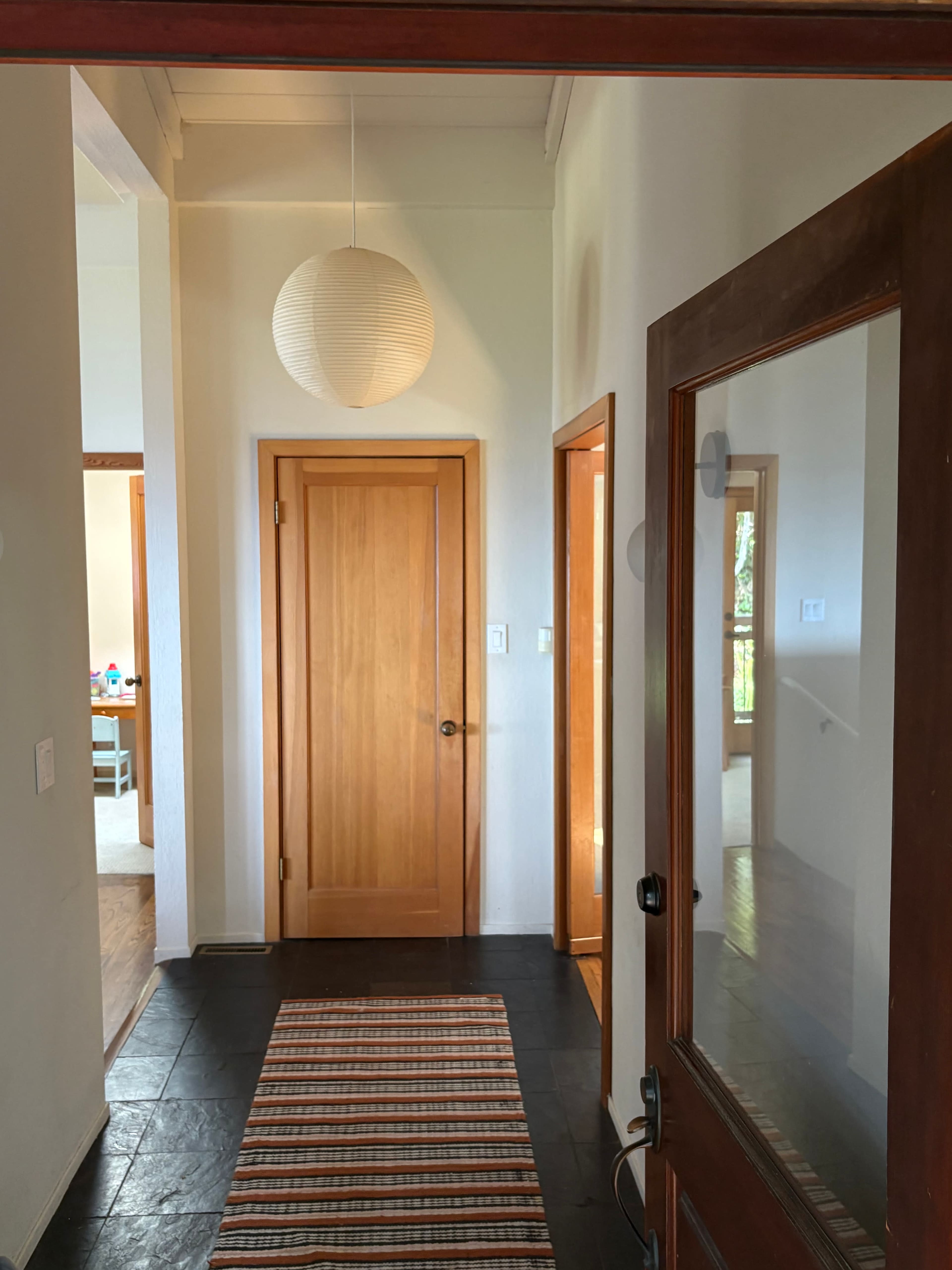 The image shows a well-lit hallway with wooden doors, a round paper lantern hanging from the ceiling, and a striped area rug on a dark floor.