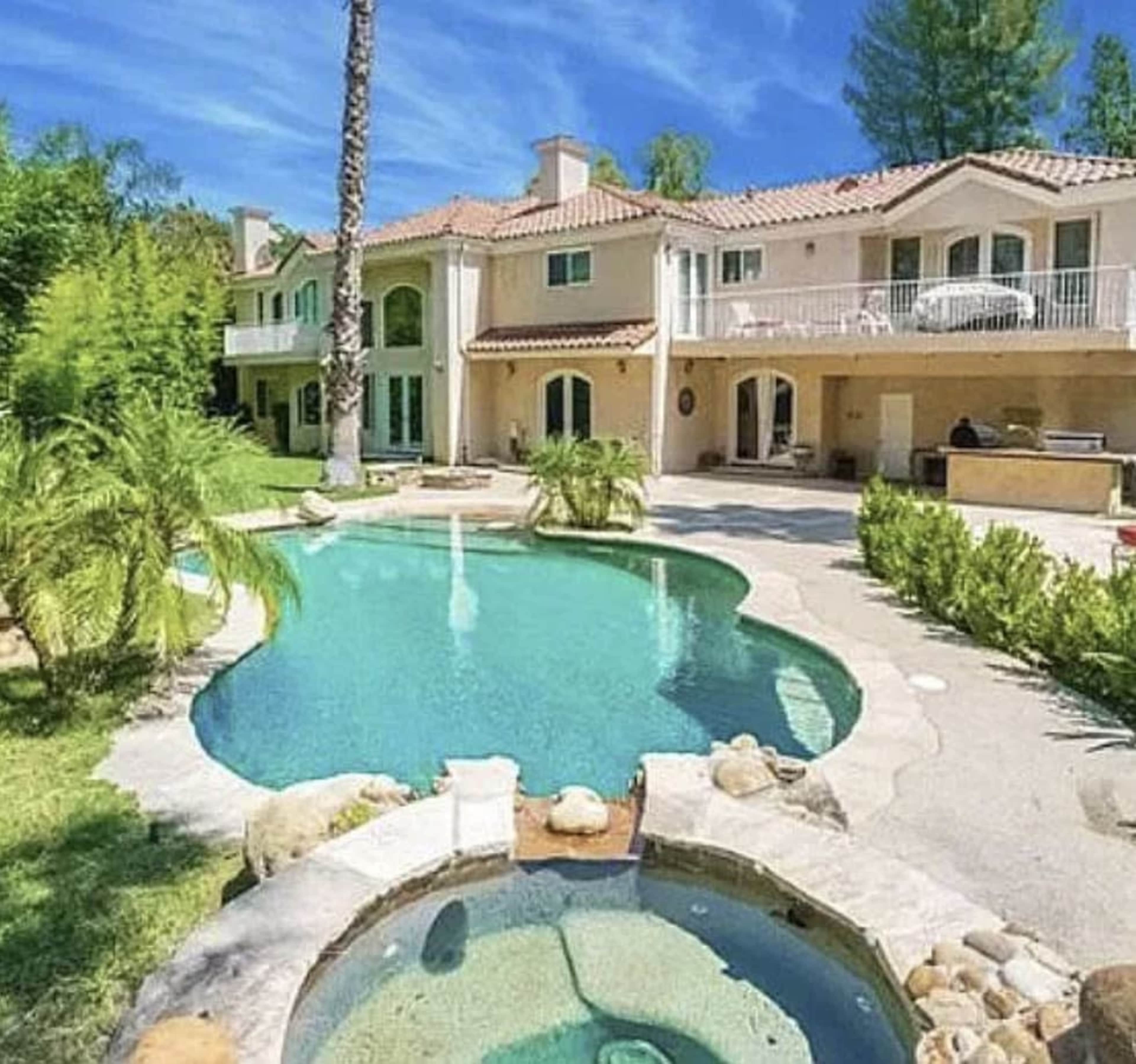 The image shows a large house with a red-tiled roof, featuring a swimming pool and a spa surrounded by lush greenery.