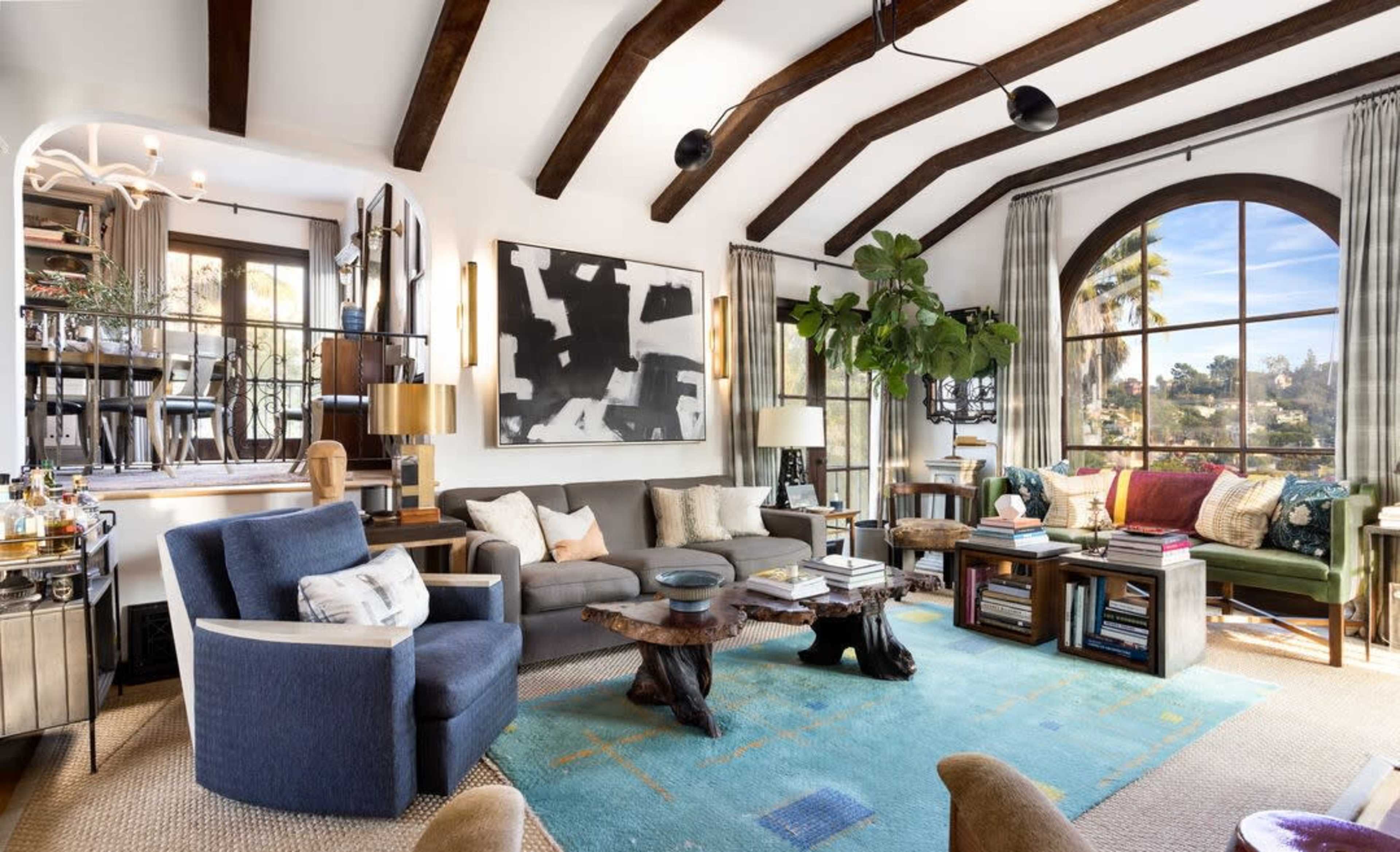 The image shows a stylish living room with vaulted wooden beams, a large window, a mix of seating options, and decorative elements like artworks and plants.