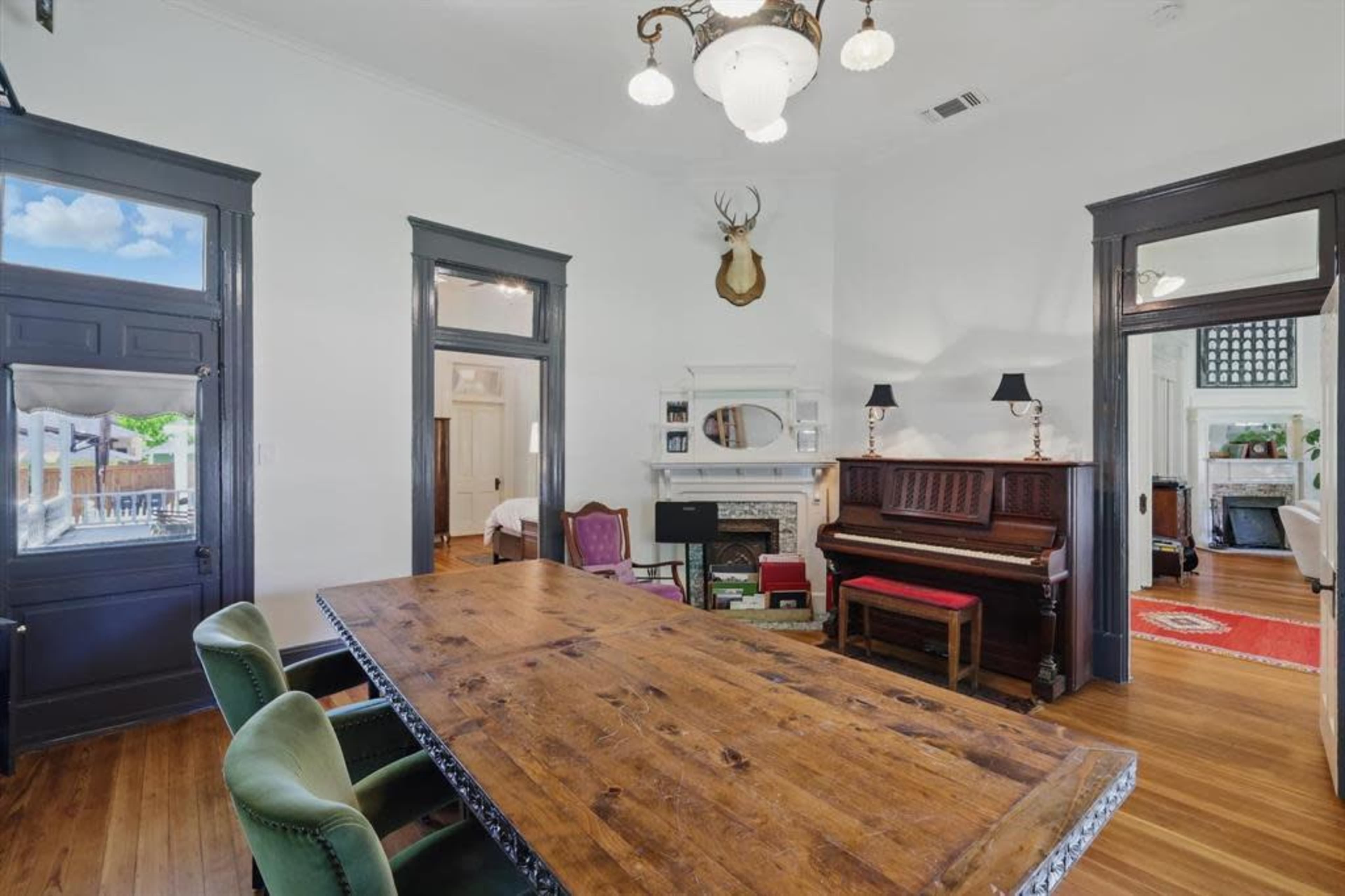 The image depicts a spacious room with a wooden dining table, a piano against the wall, and decorative accents including a mounted deer head and classic light fixtures.