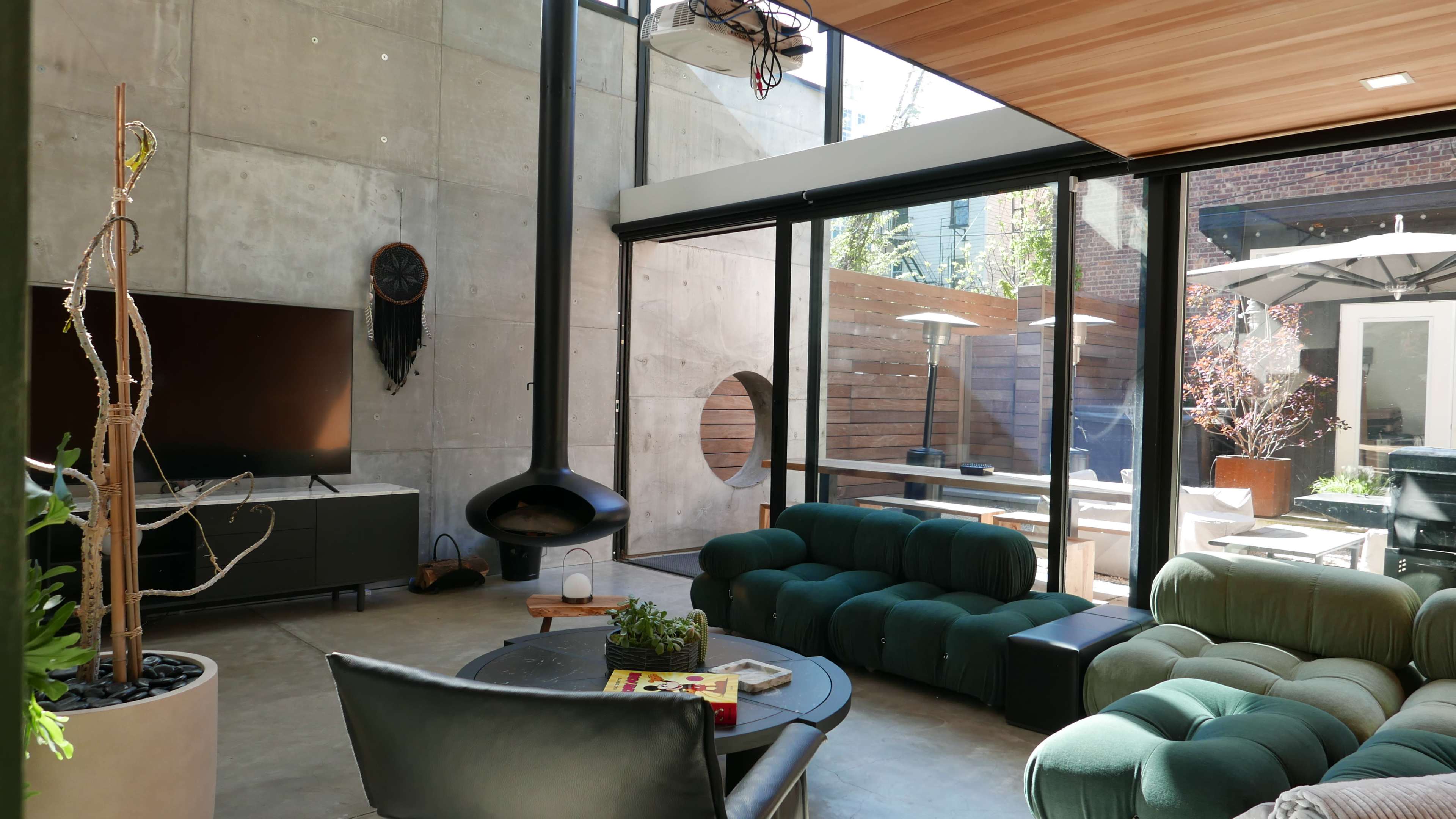 The living room features a modern design with a green sofa, a black TV stand, a circular fireplace, and large windows opening to an outdoor patio.