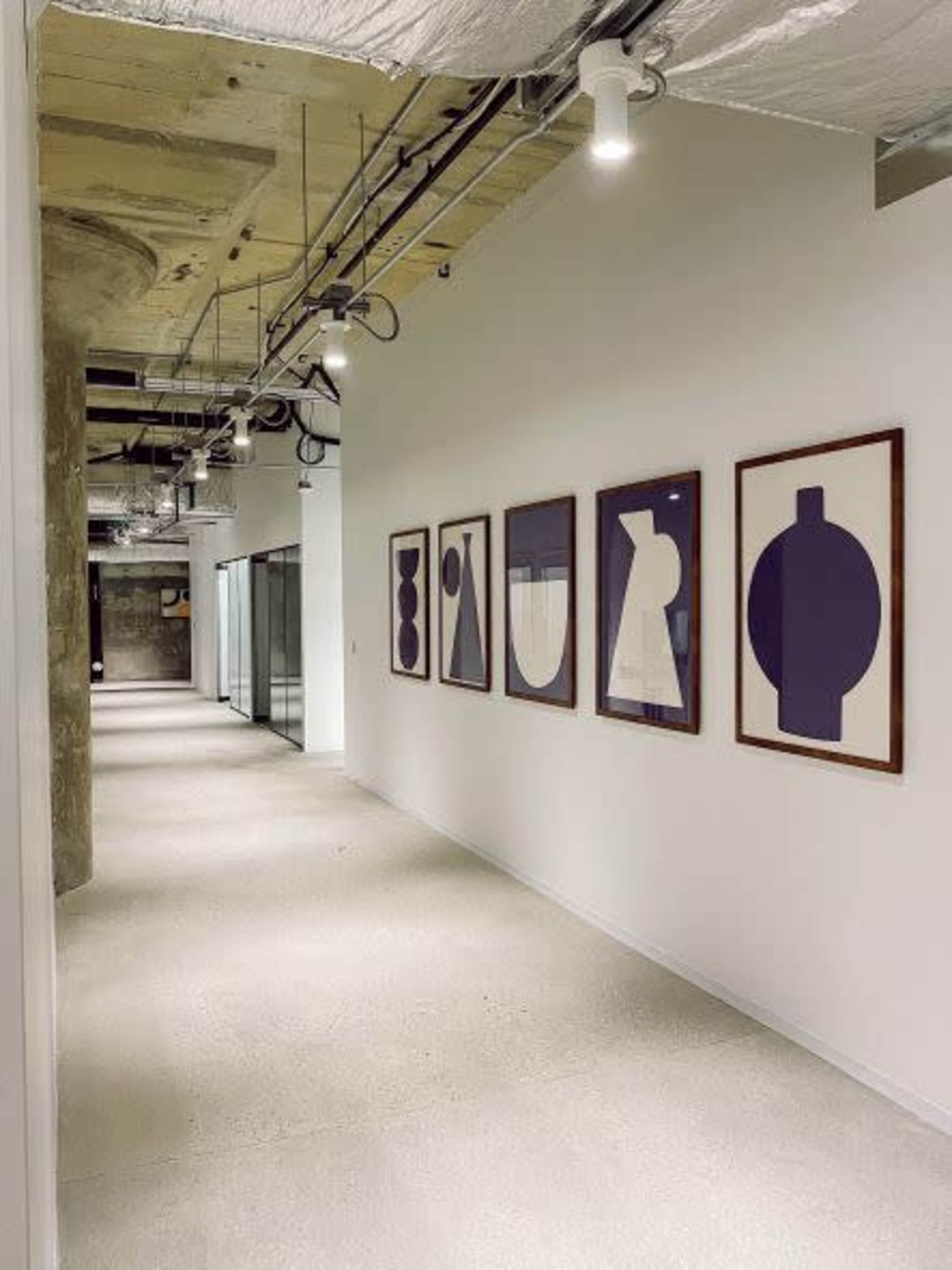A long hallway with concrete ceilings and white walls, featuring a series of framed abstract artworks on one side.