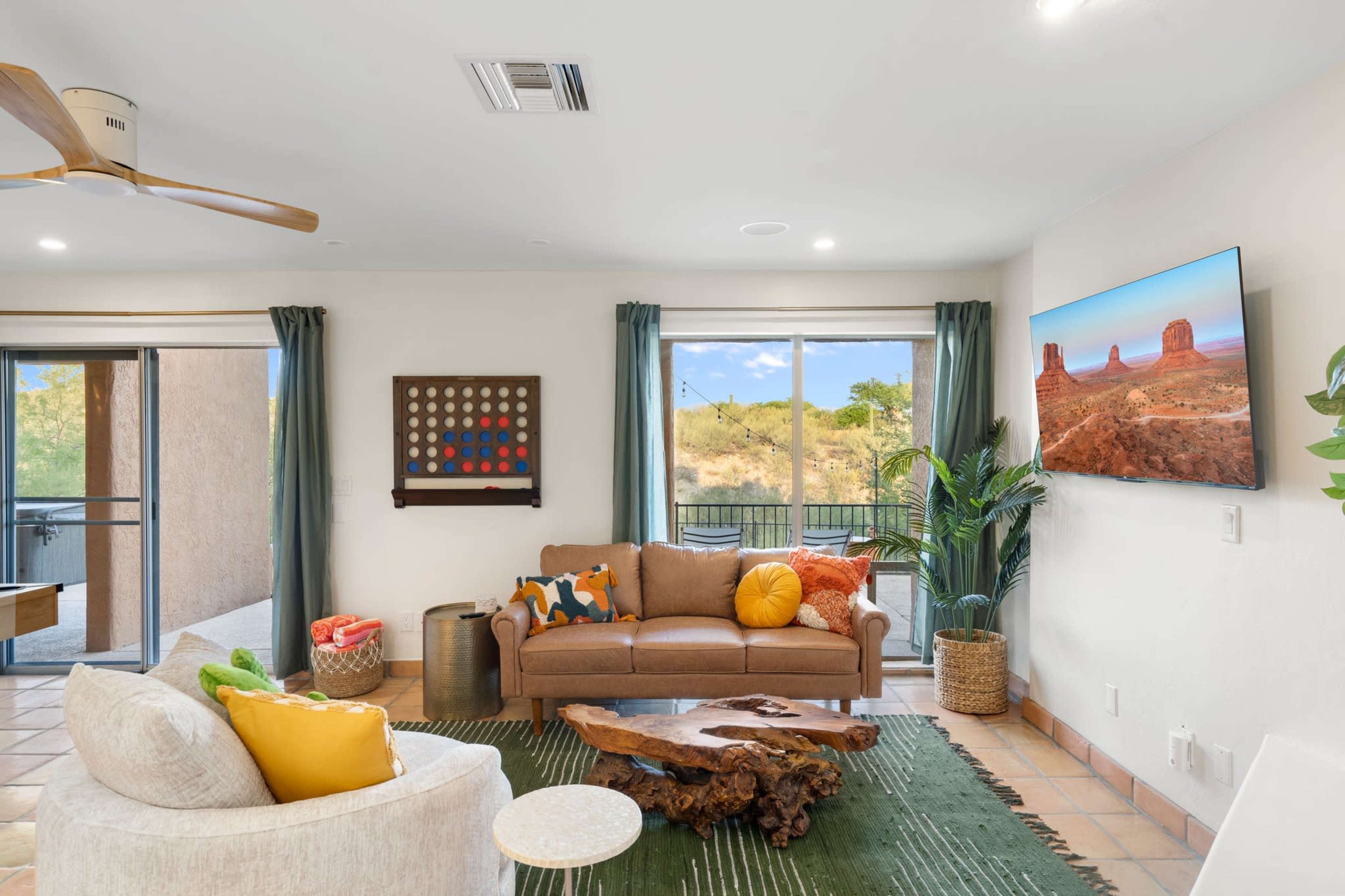 The living room features a brown leather sofa, a green area rug, a wooden coffee table, and a large wall-mounted television displaying a desert landscape.