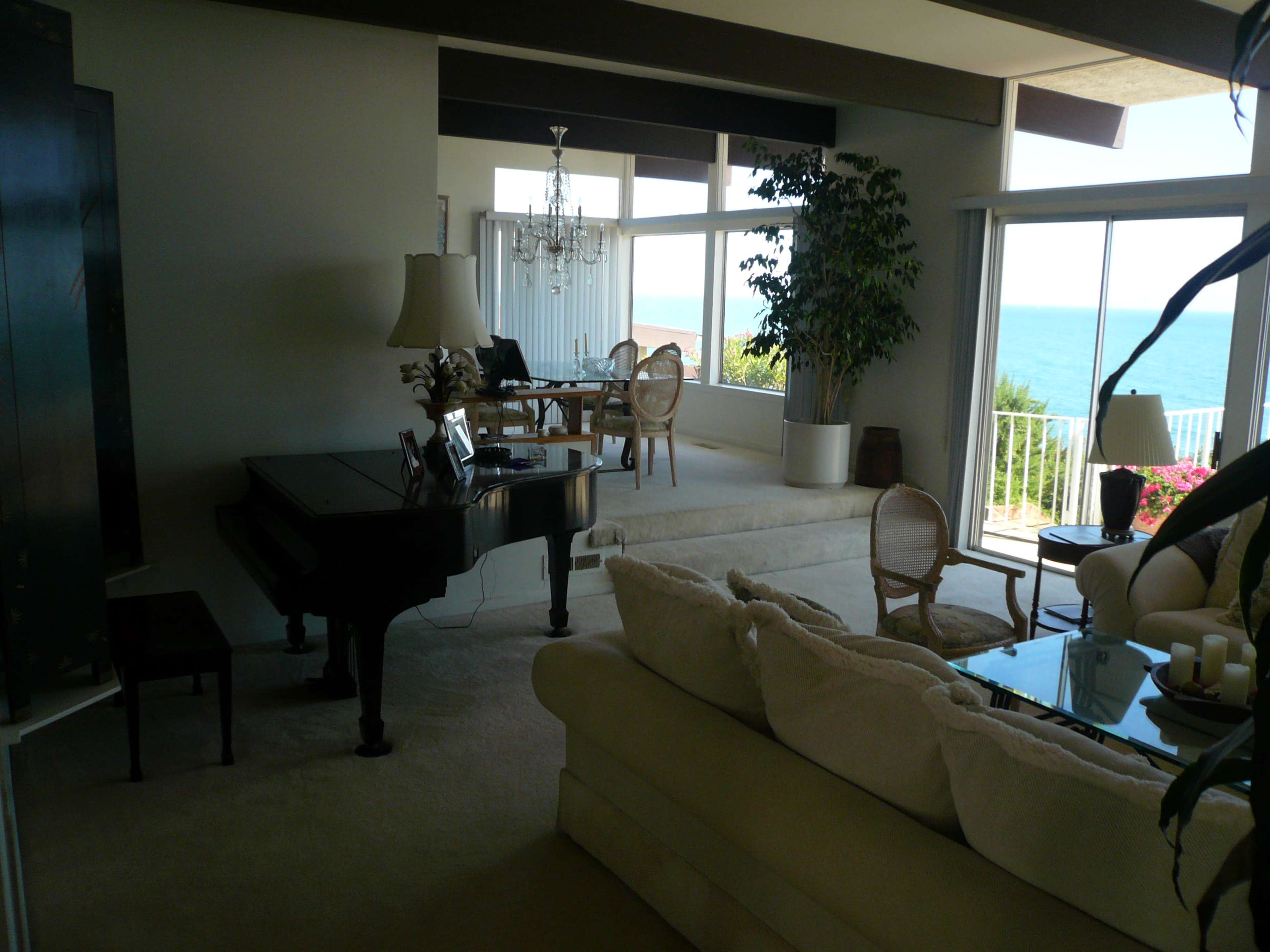 The image shows a spacious living room with a large window overlooking the ocean, featuring a grand piano, a comfortable sofa, and a dining area in the background.