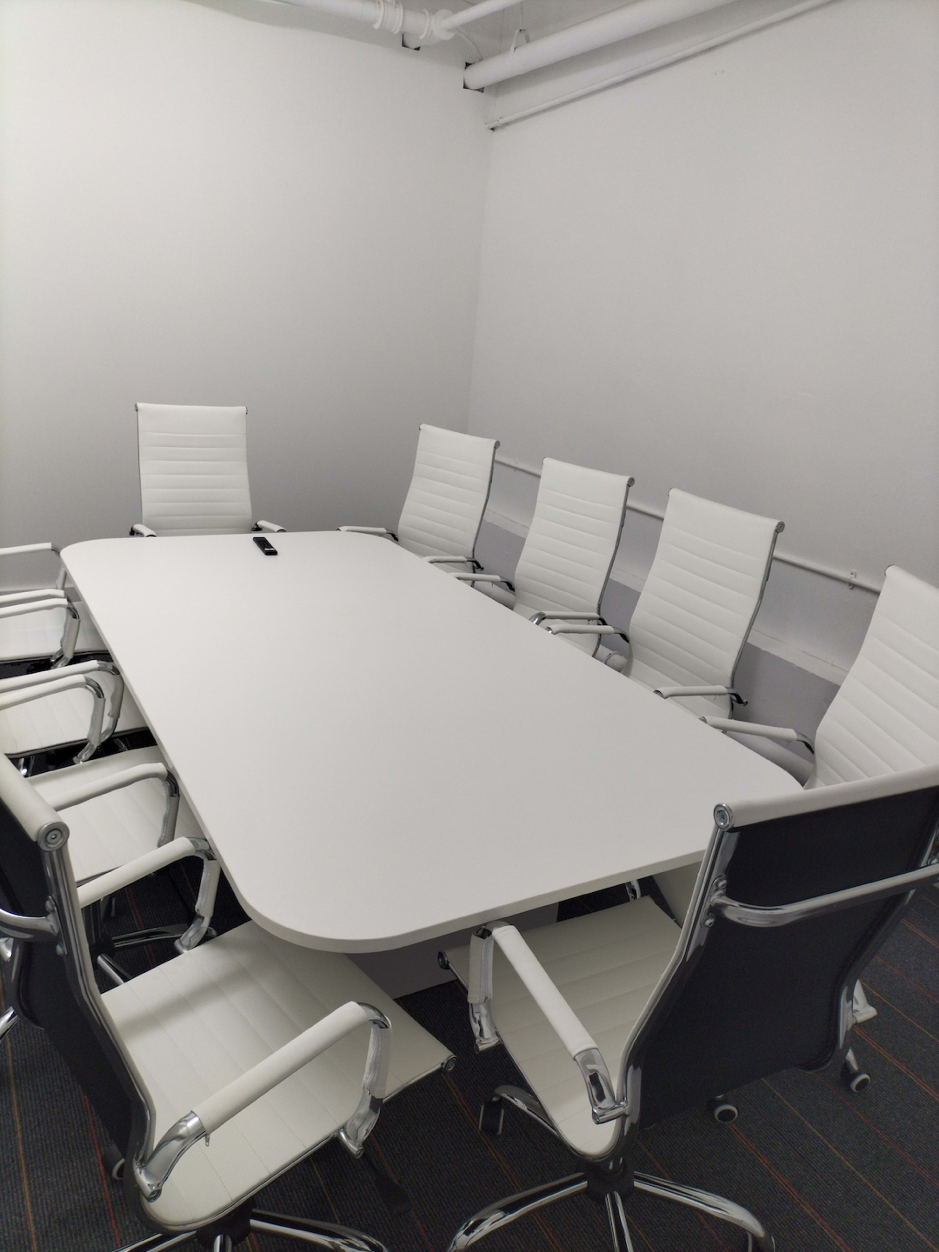 A large, modern conference table surrounded by eight white chairs is set up in a brightly lit meeting room.