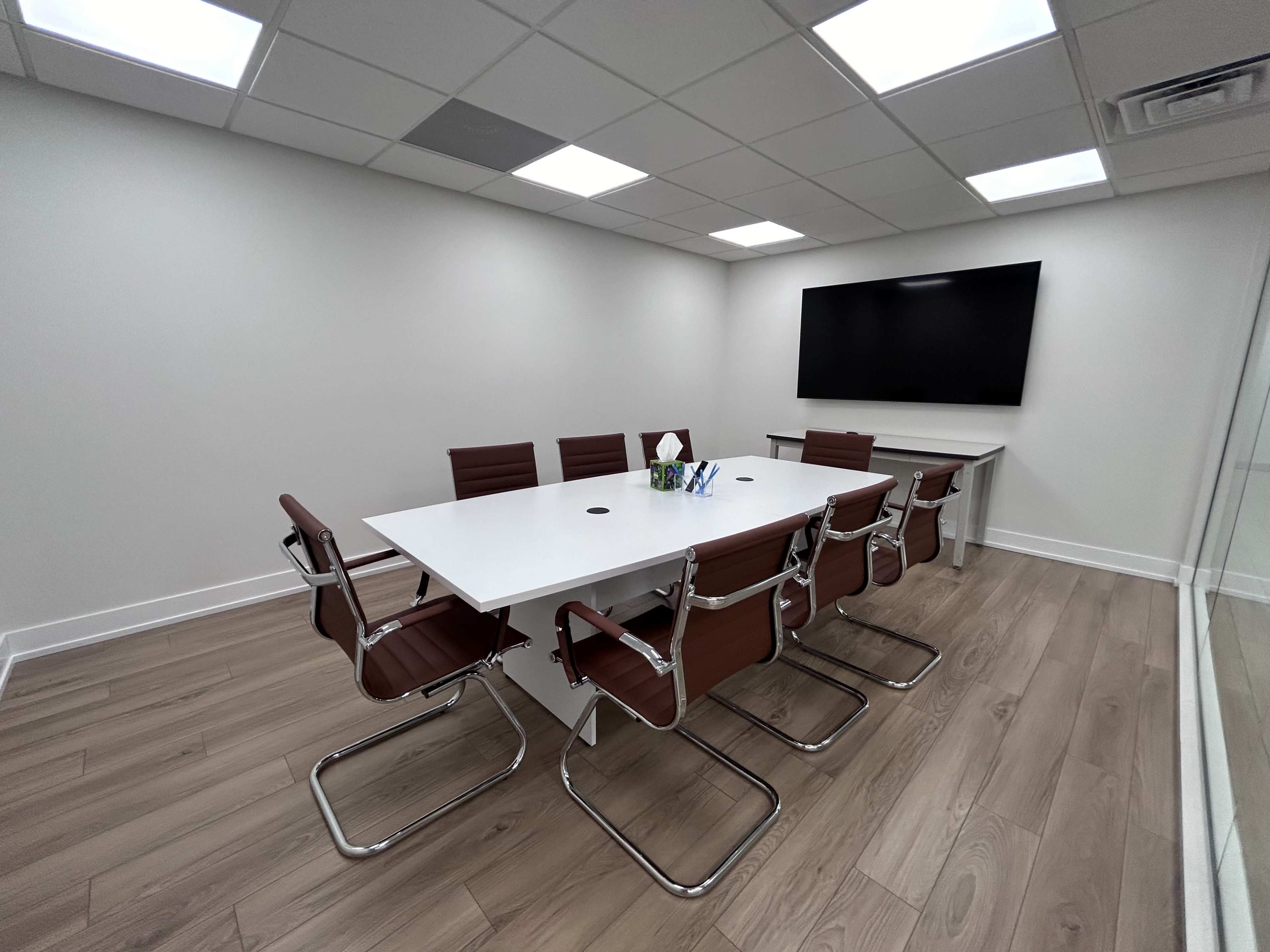 A modern conference room features a white table surrounded by brown leather chairs, with a large screen mounted on the wall.