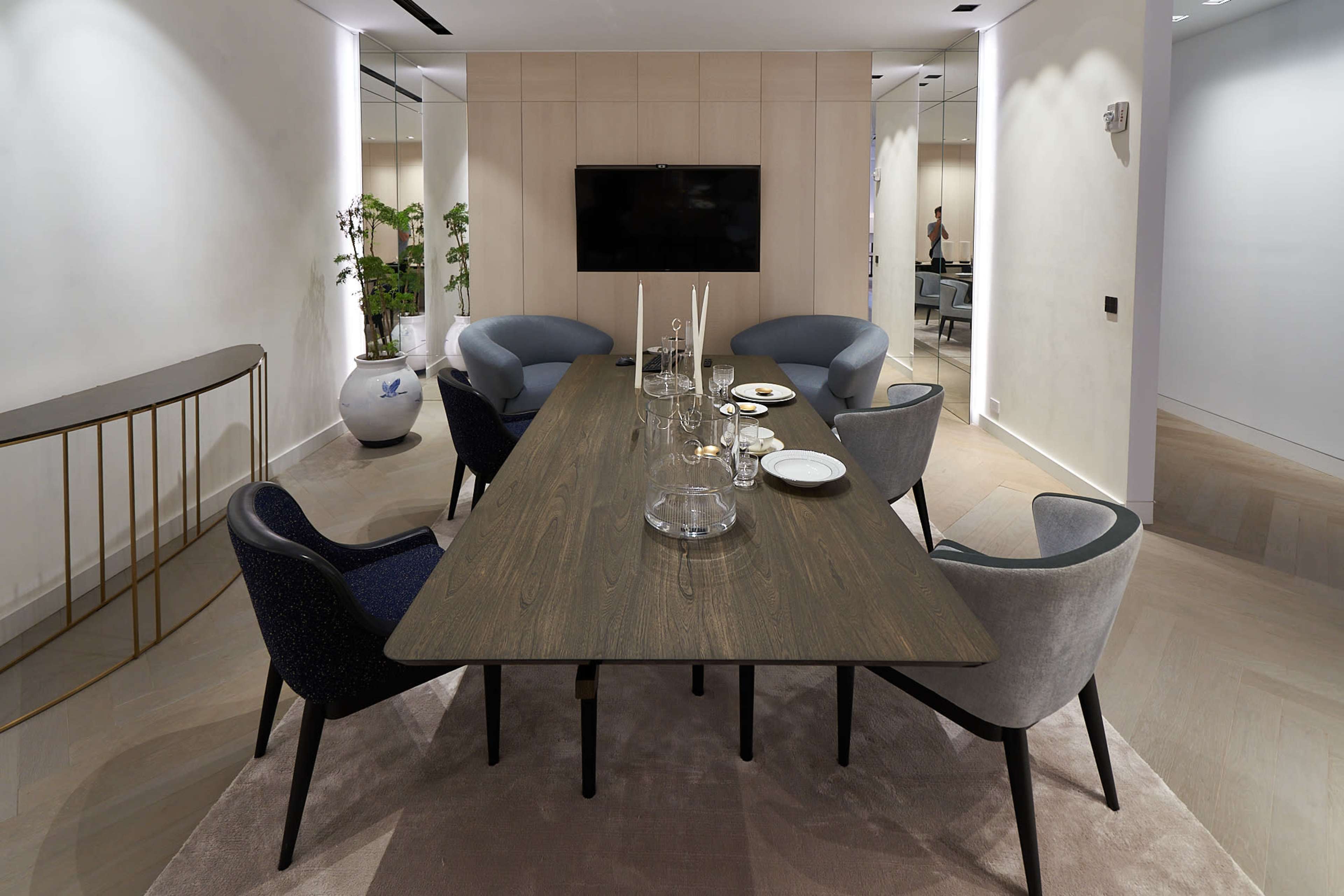 A modern dining area features a long wooden table surrounded by upholstered chairs, with a television mounted on the wall and decorative plants in the background.