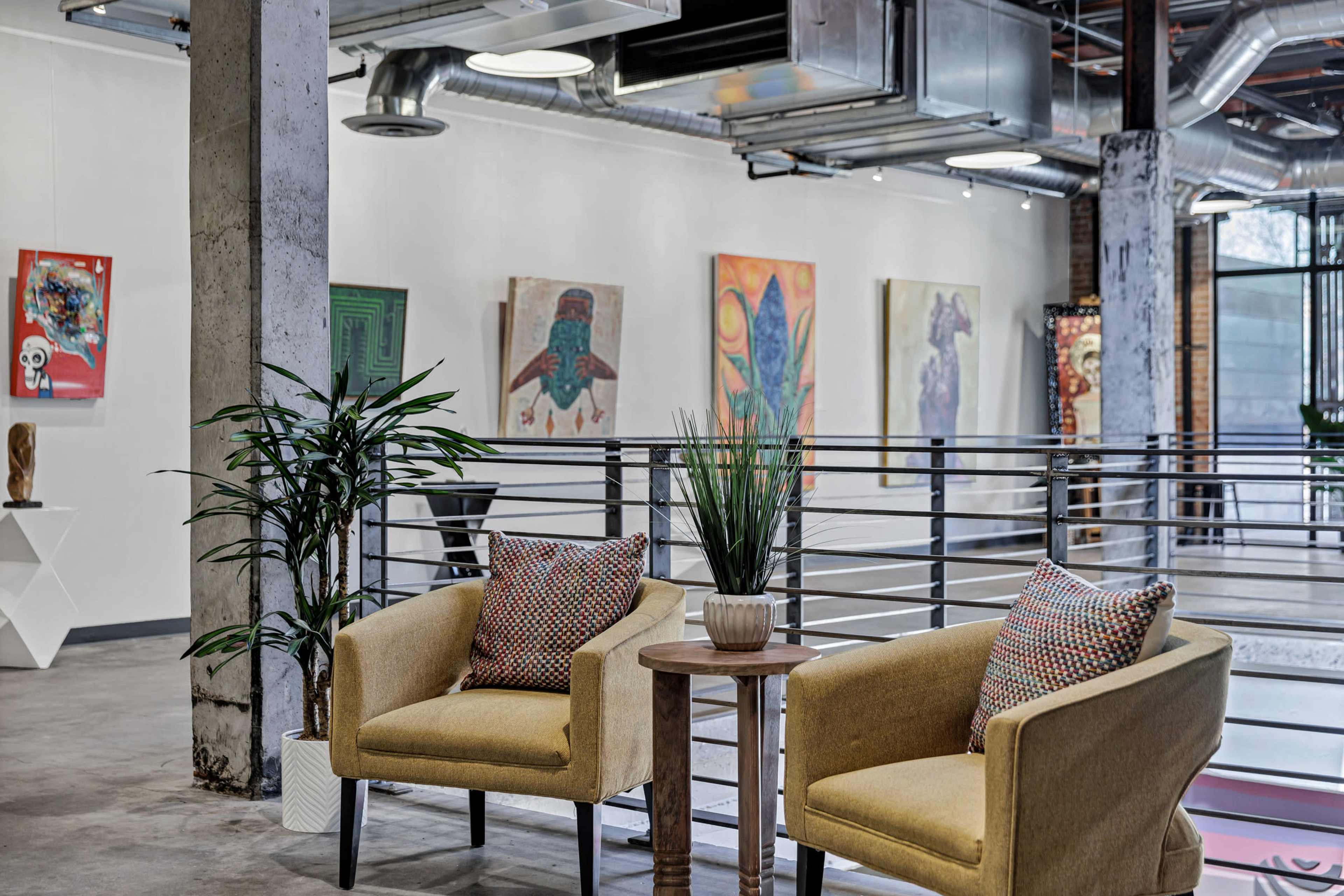 The image depicts a modern gallery space featuring two yellow armchairs with patterned cushions, a small wooden table between them, and colorful artwork hanging on the walls.