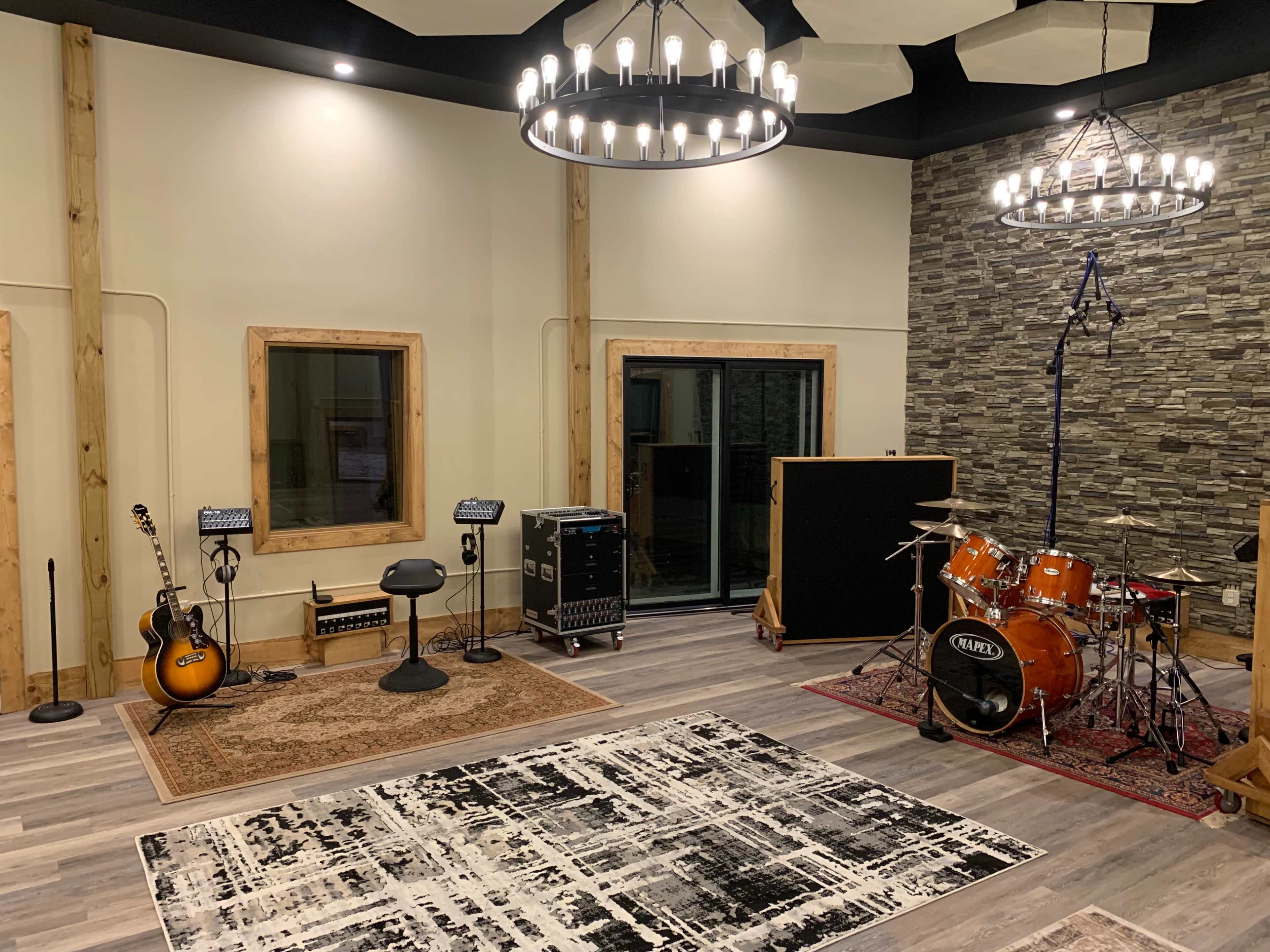 The image shows a music studio featuring a drum set, electric guitar, sound equipment, and acoustic treatment on the walls.