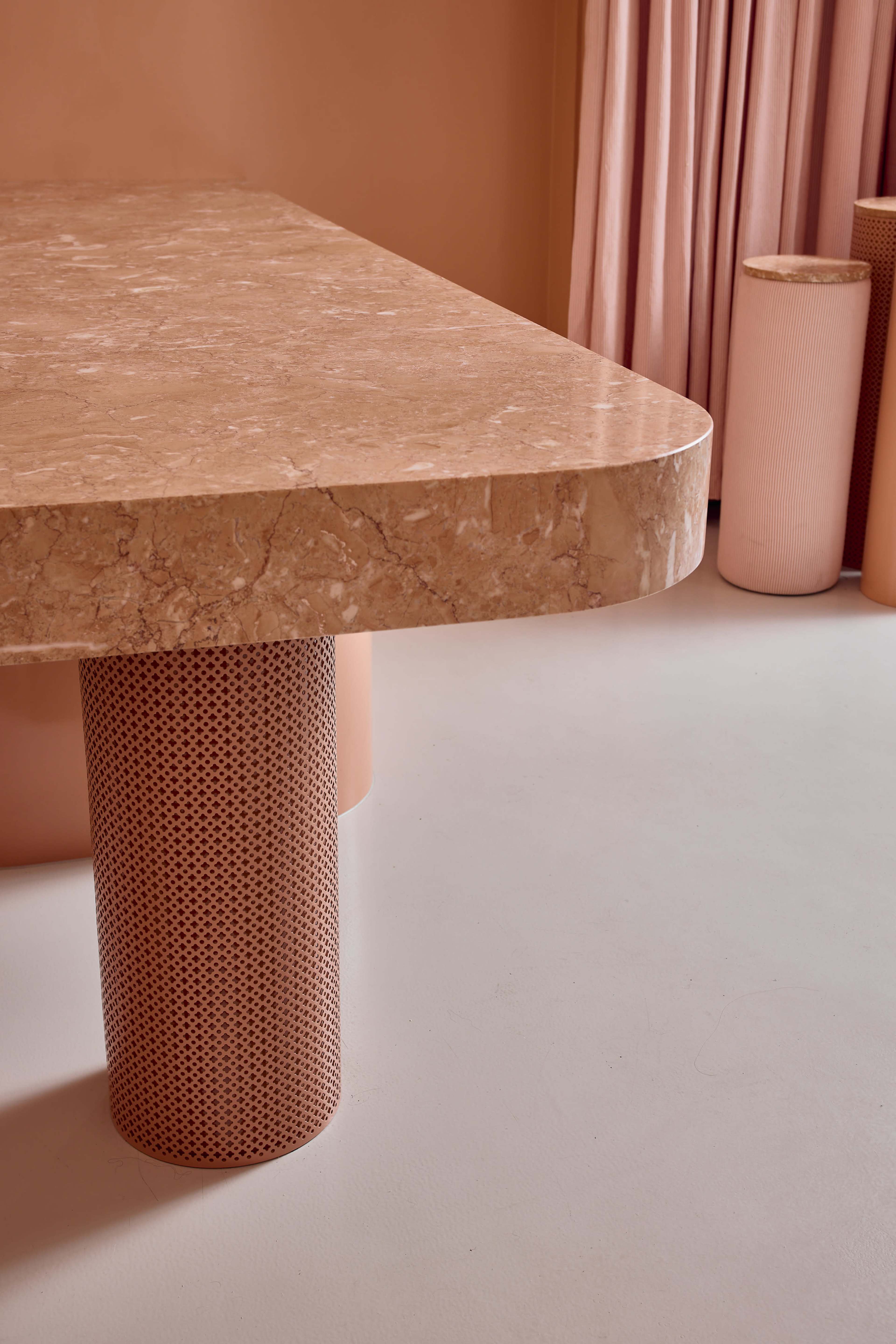 A marble table with a rounded edge rests on a cylindrical, perforated pink column, set against a backdrop of soft pink curtains and decorative cylindrical elements.