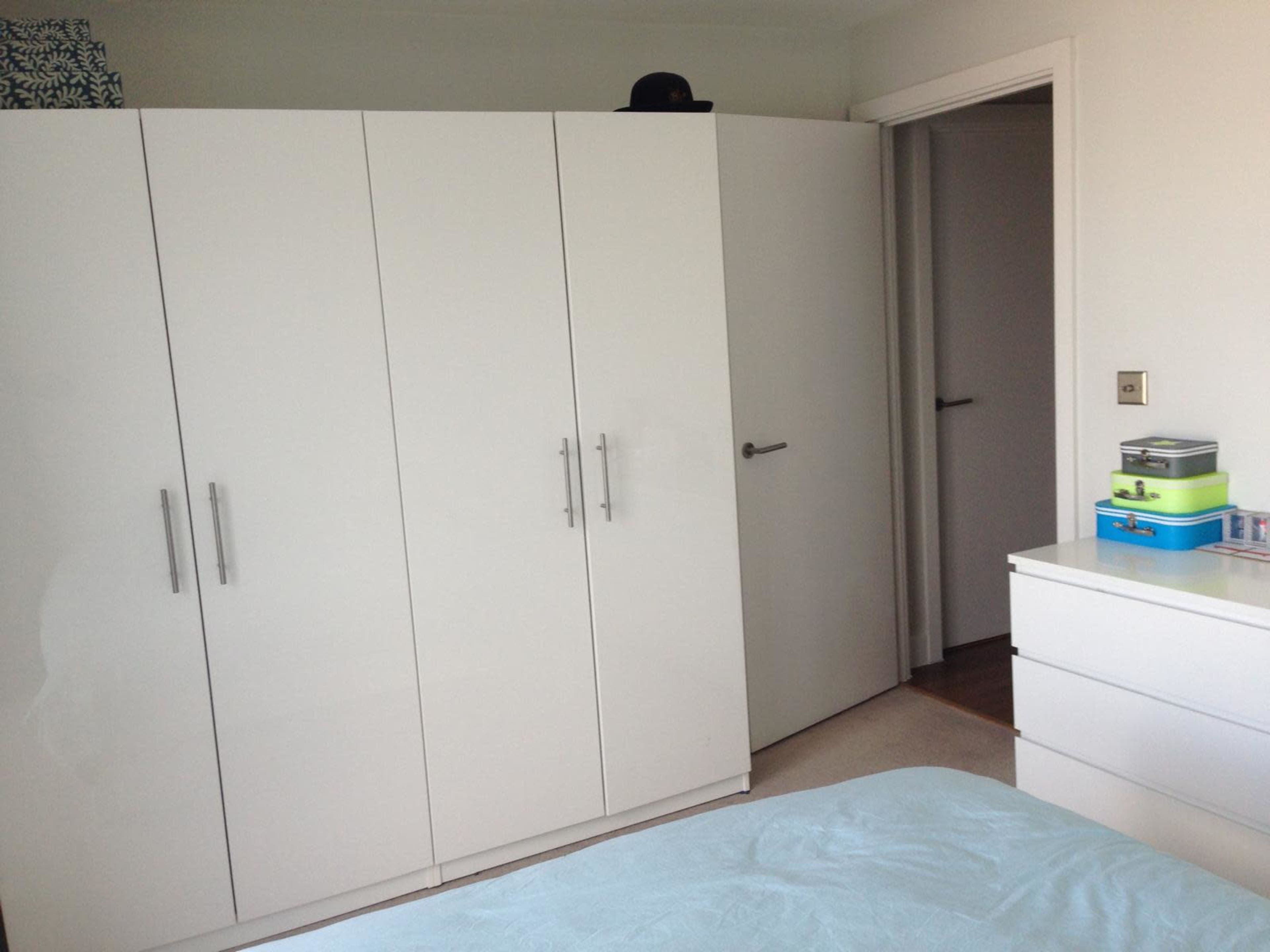 The image shows a modern bedroom with a white wardrobe, a closed door leading to another room, and a dresser with colorful storage boxes on top.