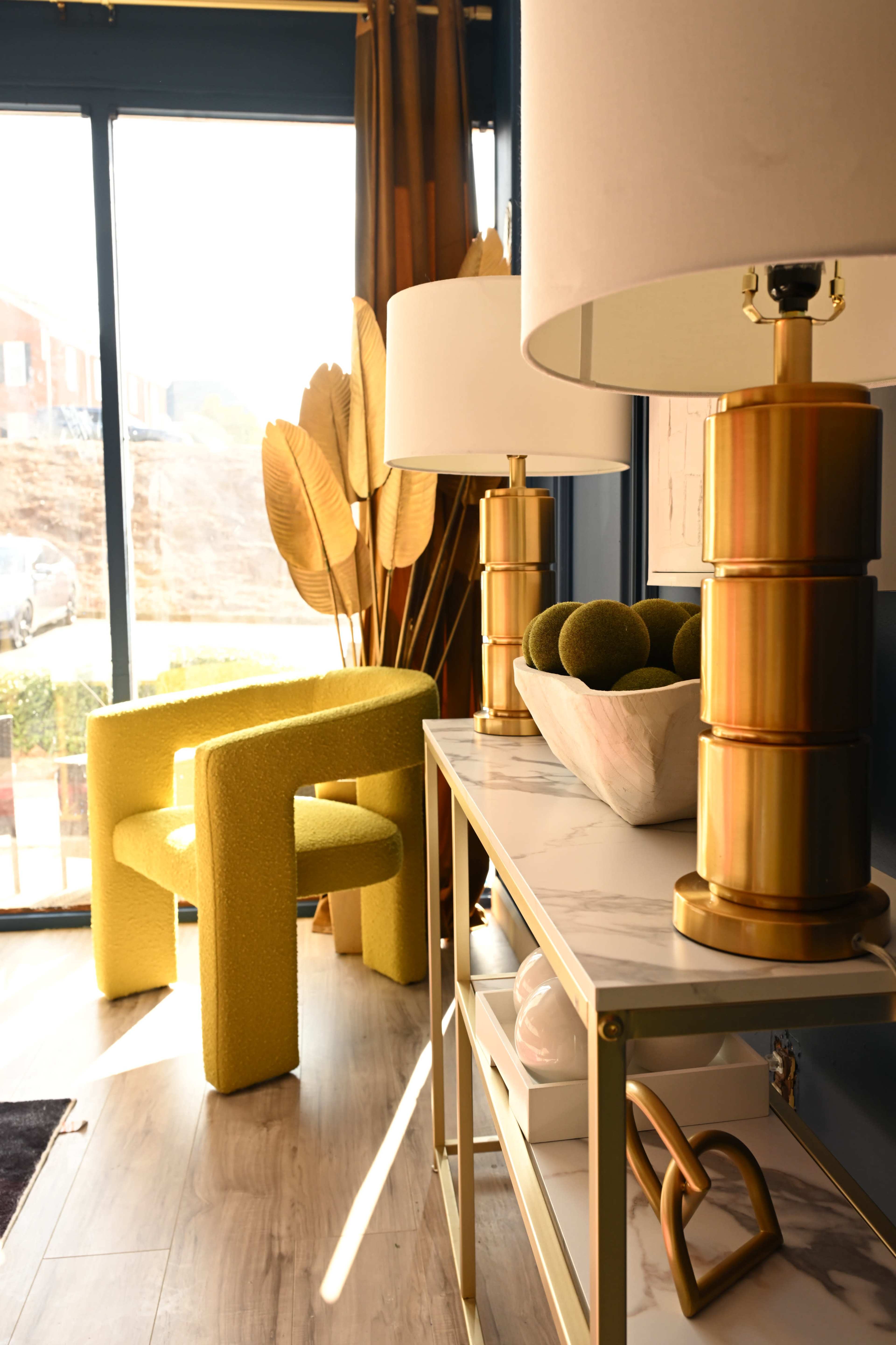 The image shows a modern living space featuring a yellow upholstered chair, a marble-top console table with decorative items, and lamps with gold bases, all positioned near a large window.