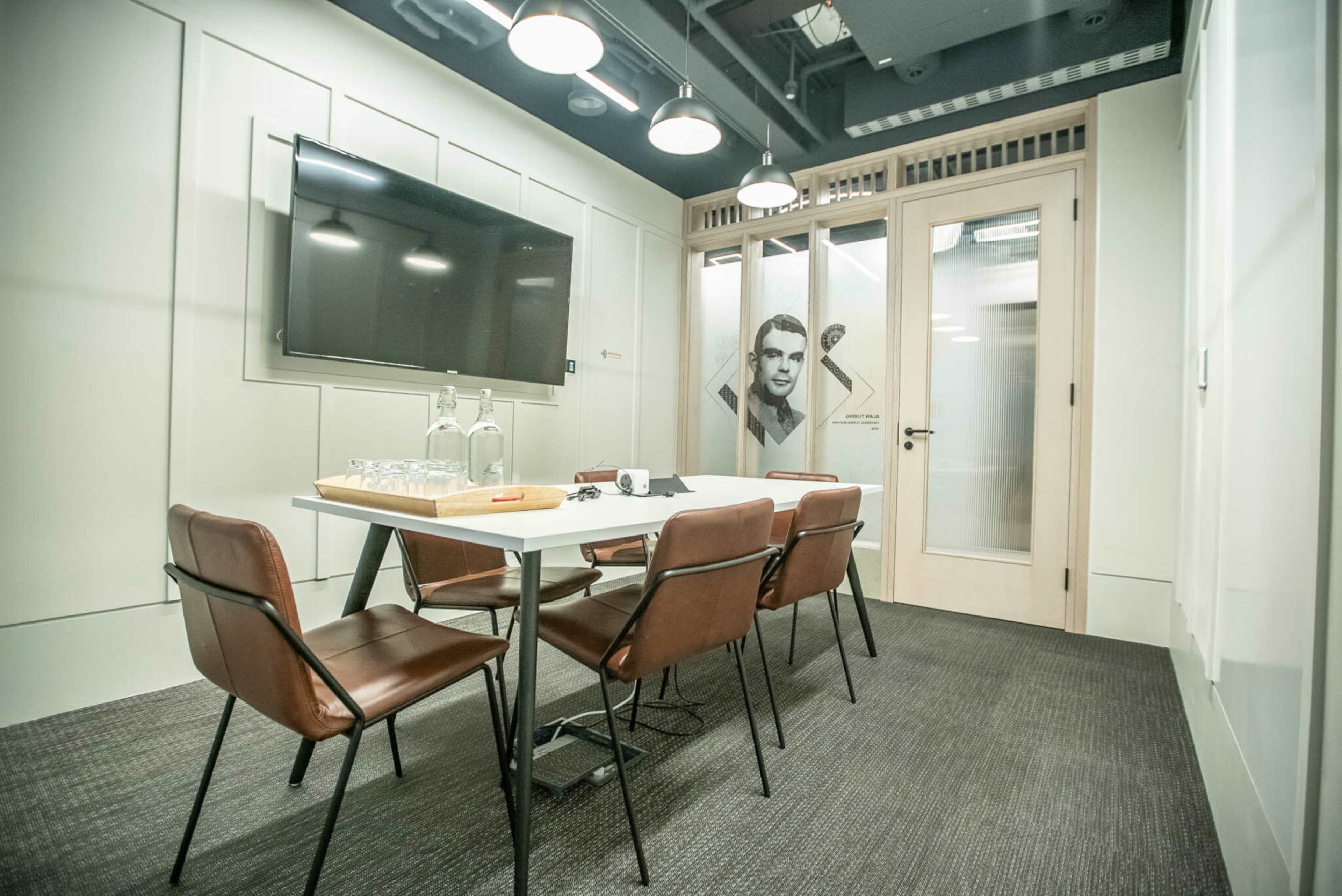 A modern conference room features a table with four chairs, a television on the wall, and glass doors displaying a mural.