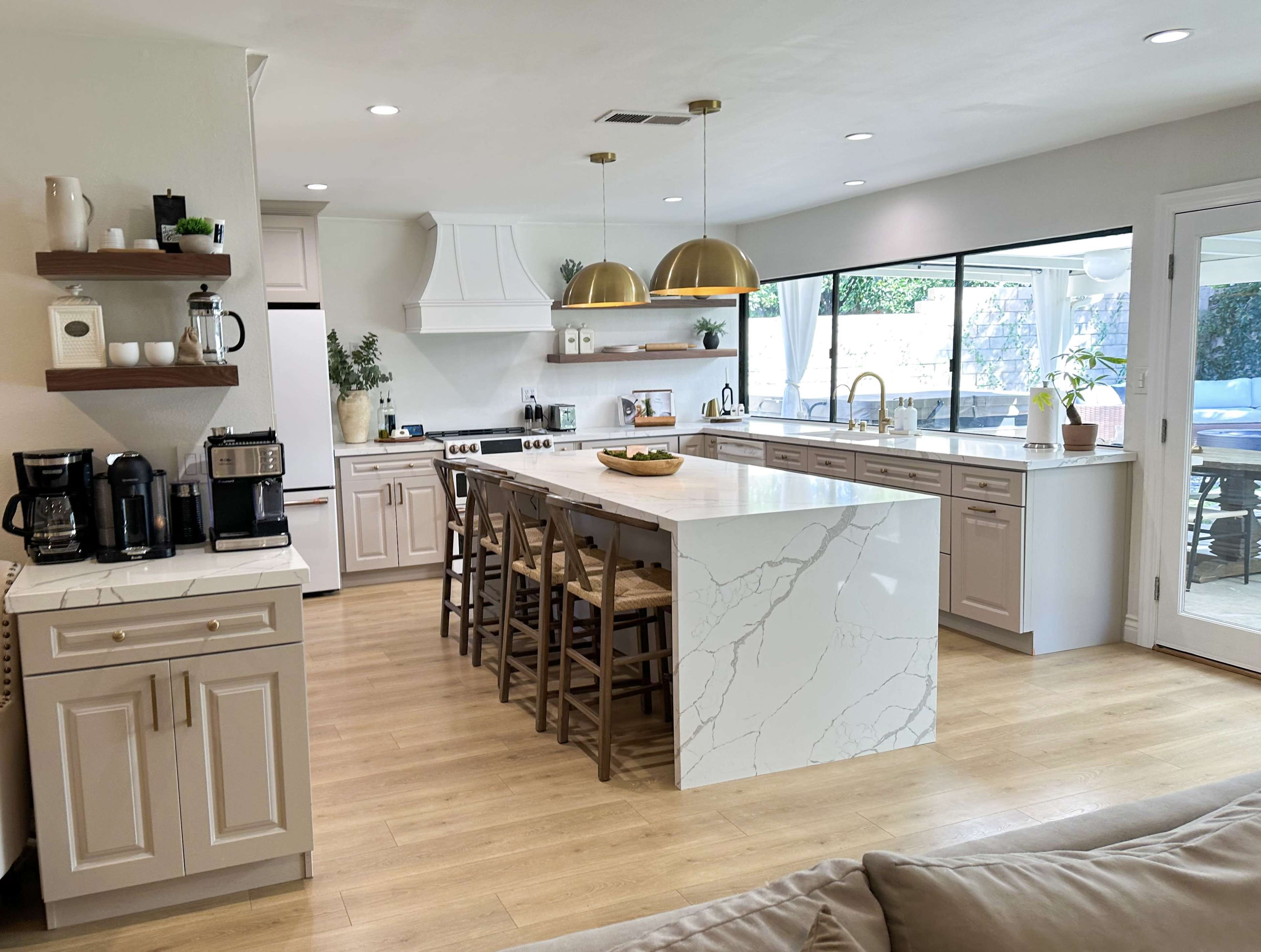 A modern kitchen features beige cabinets, a large island with white marble countertop, pendant lights, and large windows overlooking an outdoor space.