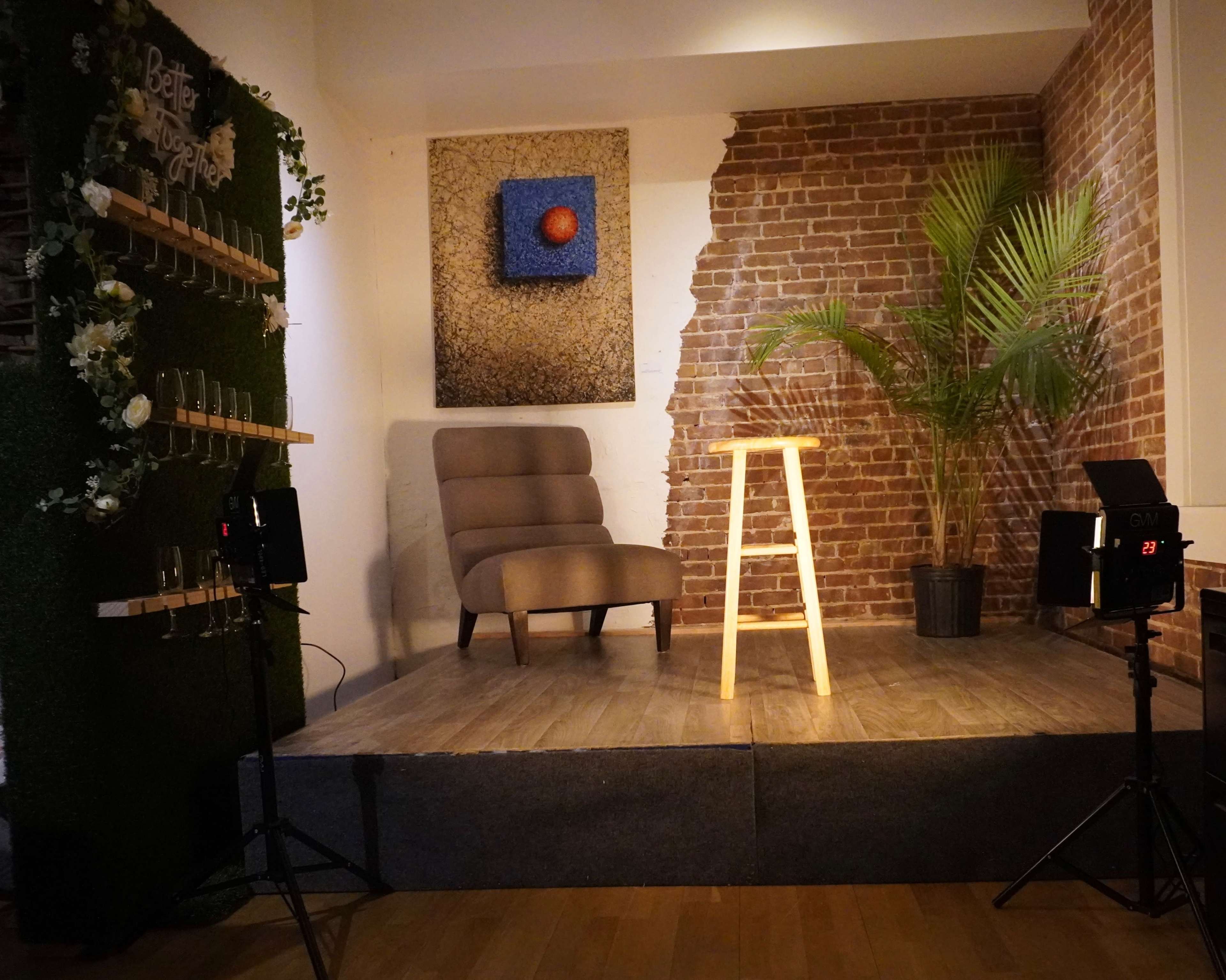 A small stage with a wooden stool, surrounded by a brick wall and a green plant, with lighting equipment positioned nearby.