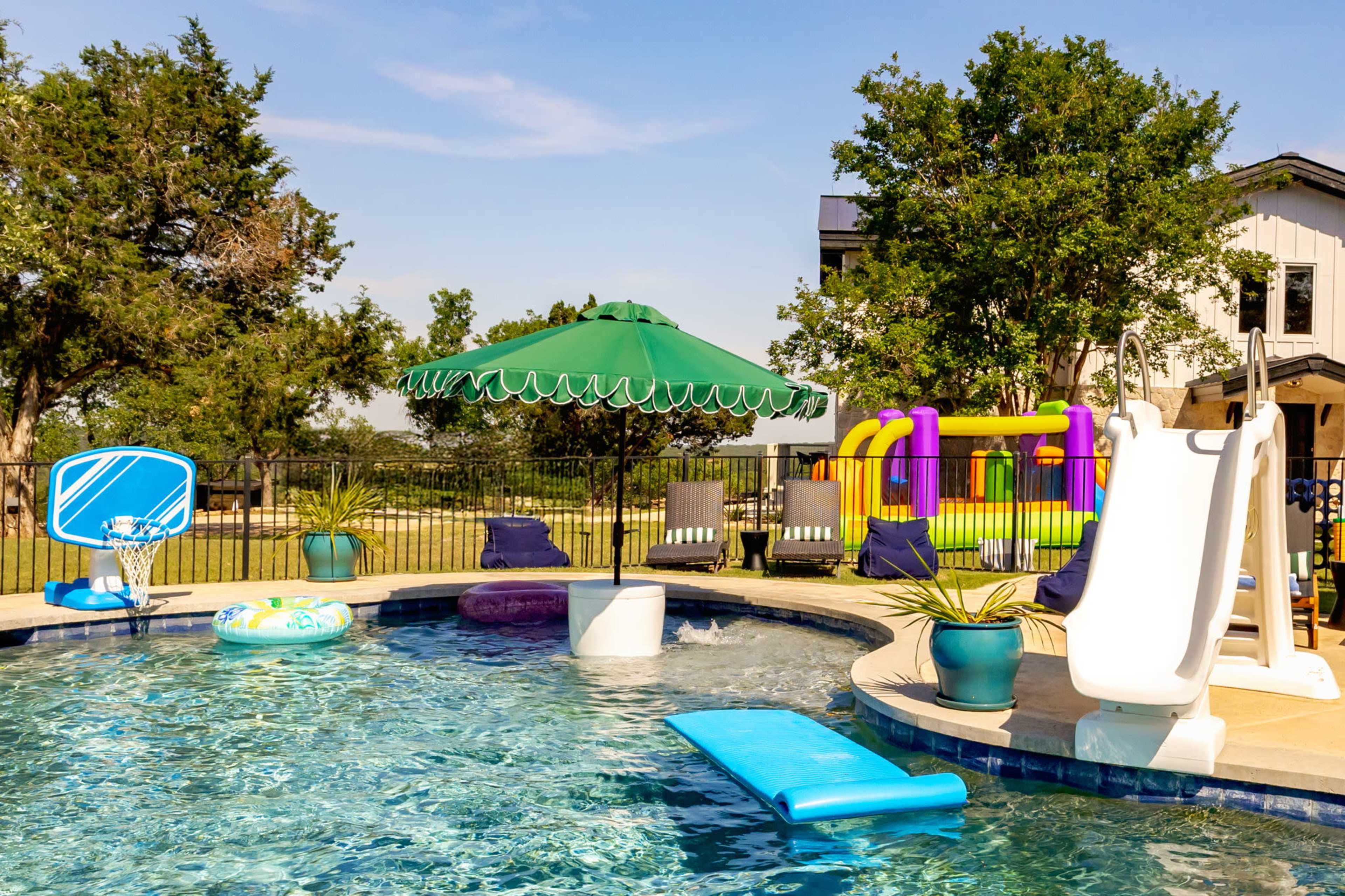 The image shows a residential backyard featuring a swimming pool with a green umbrella, pool floats, a slide, and lounge chairs under bright blue skies.