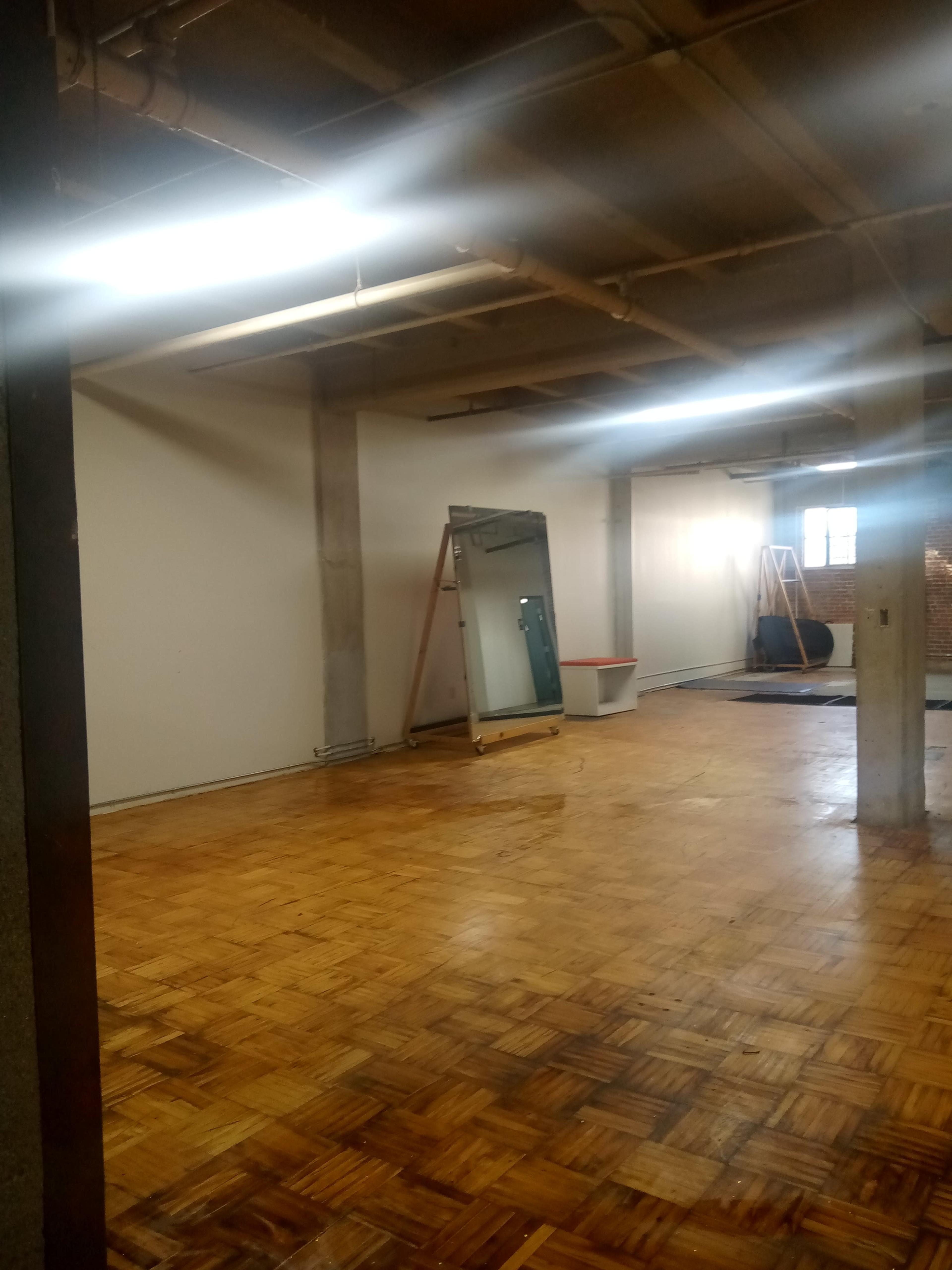 An empty room with a wooden floor, a large mirror leaning against the wall, and a white bathtub in the background.