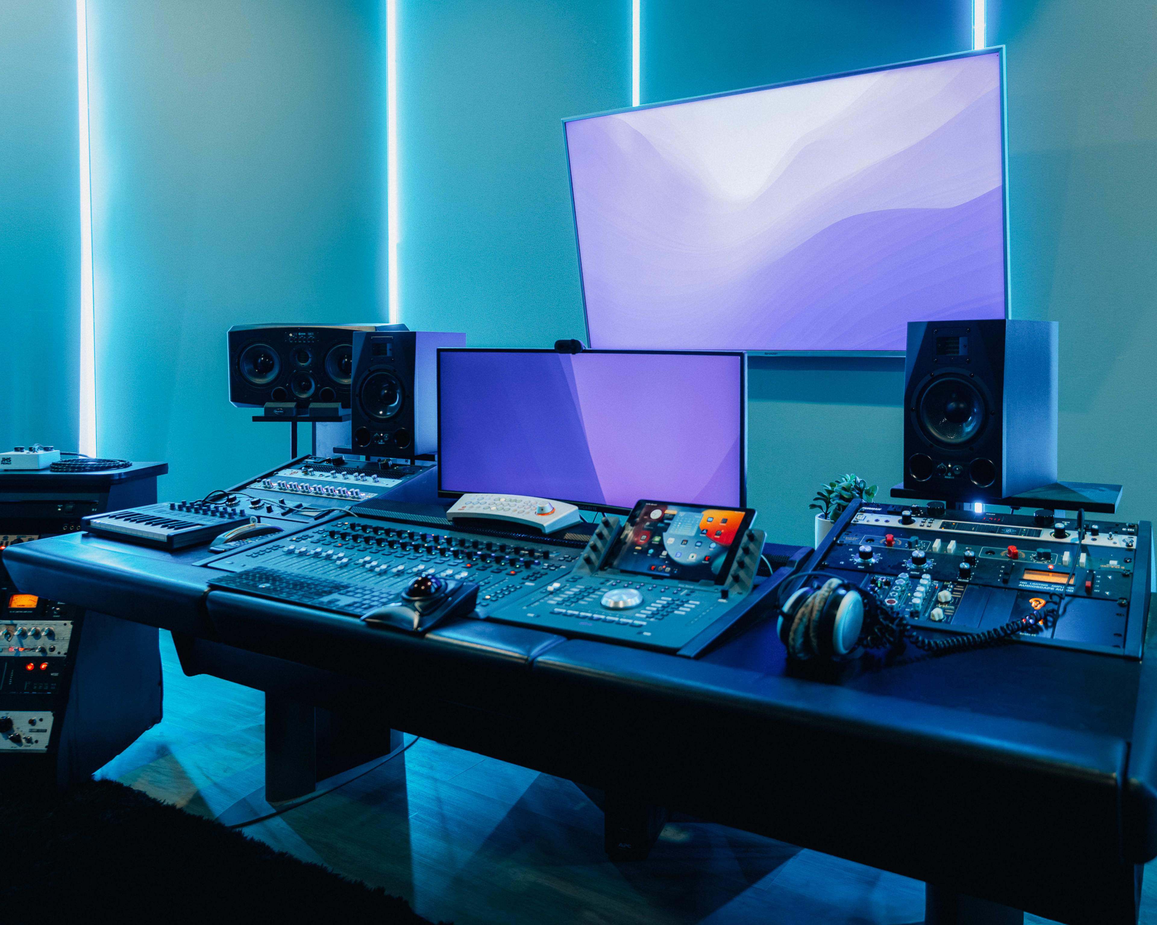 The image shows a modern music studio with a large sound mixing console, multiple monitors, and speakers, all set against a backdrop of blue LED lighting.