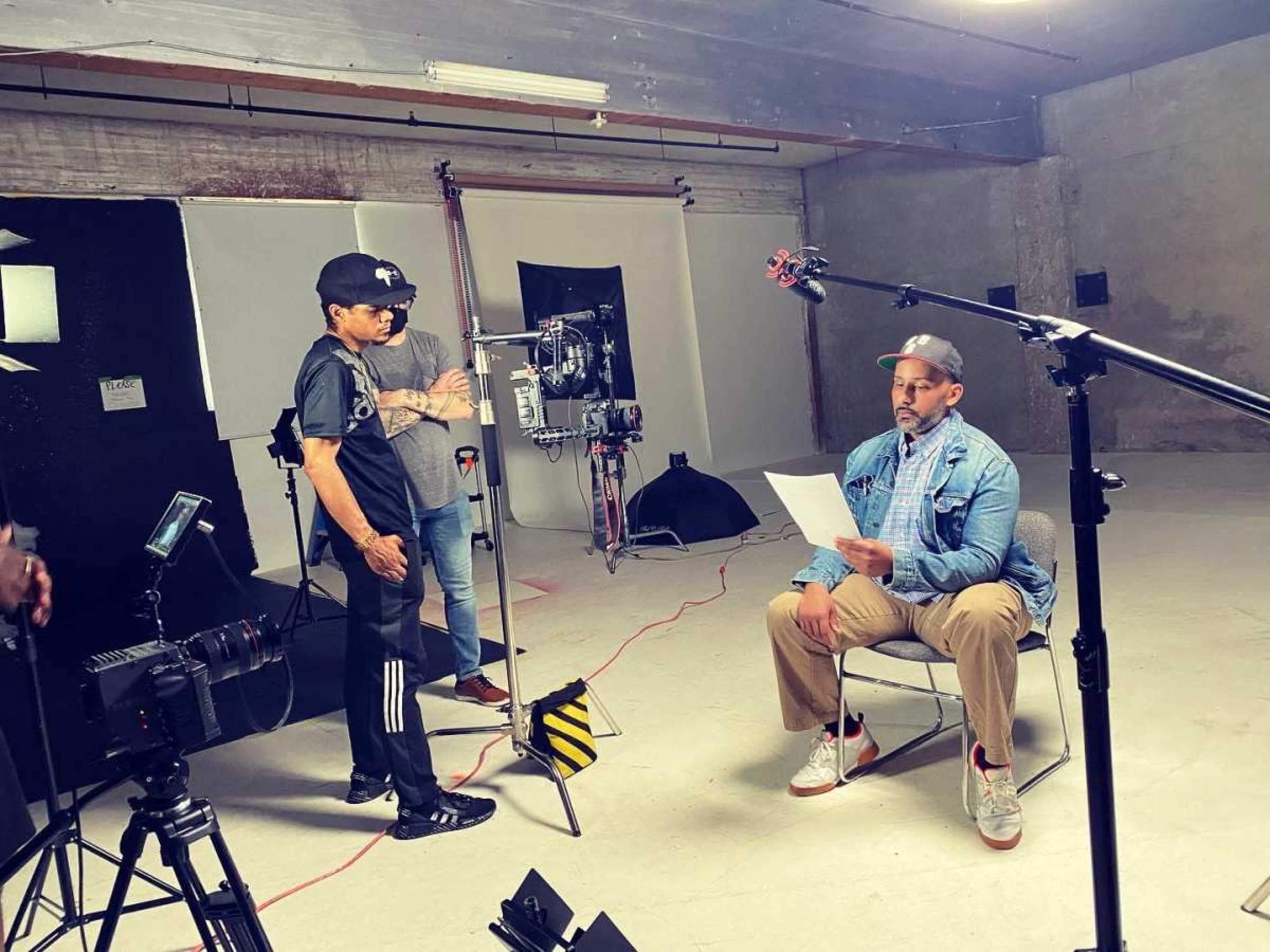 A man sits on a chair reading a script in a video production studio while another person adjusts equipment nearby.