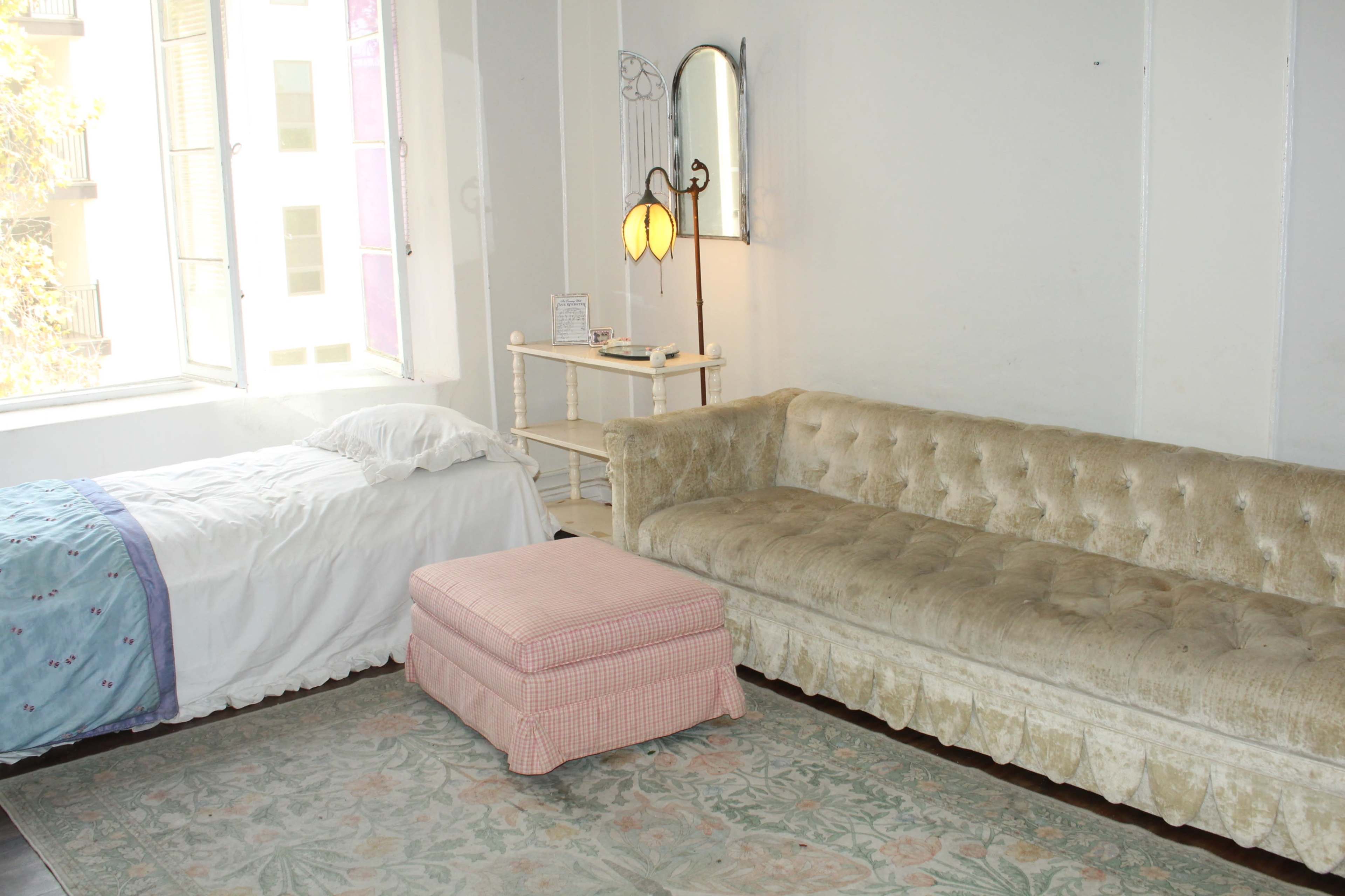 The image shows a room with a bed covered in white linens, a vintage couch, an ottoman, and a small table with a lamp, all situated near a window.