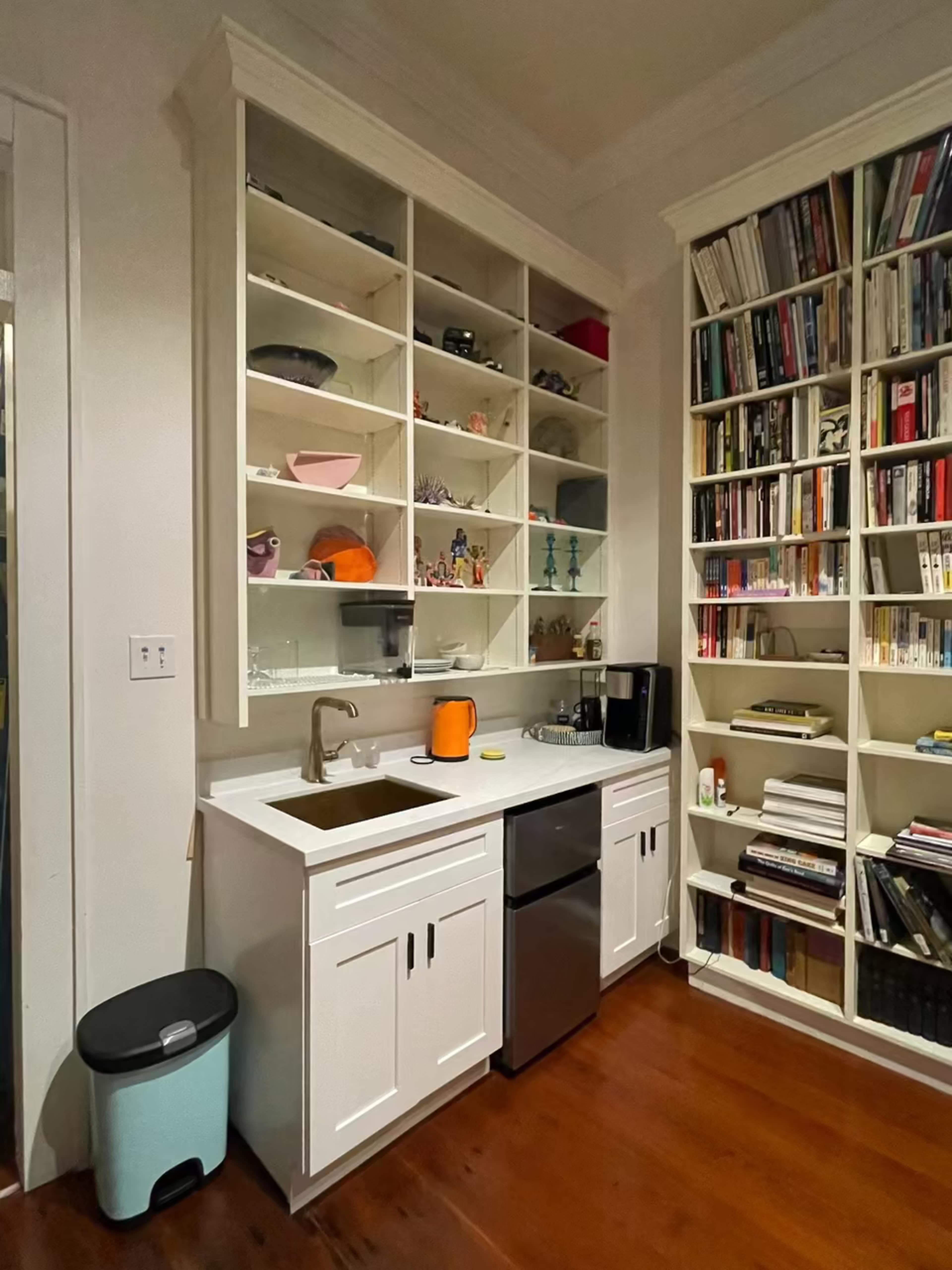 The image shows a small kitchen area featuring a sink, a refrigerator, and a bookshelf filled with books and decorative items.