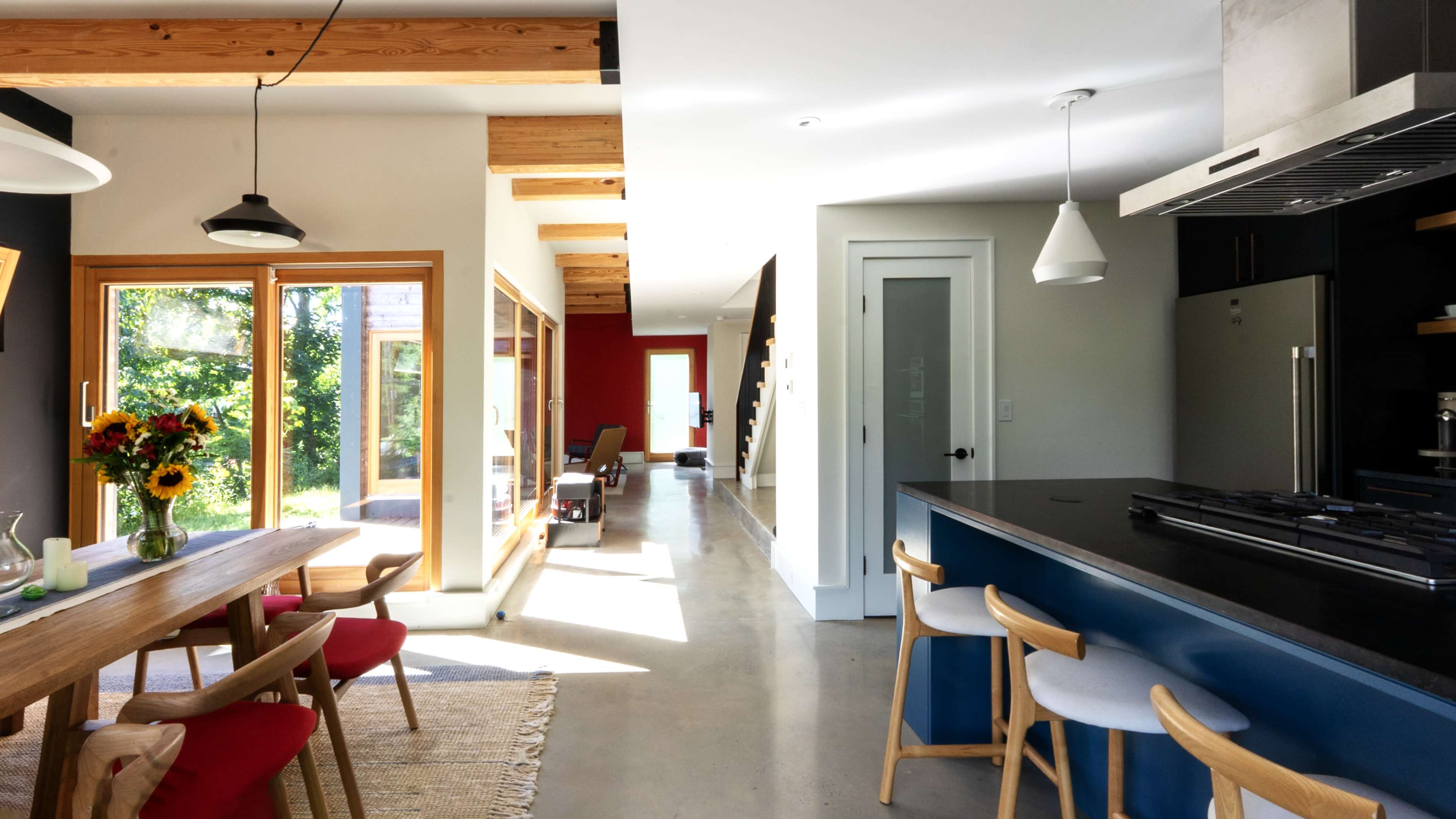 The image shows a modern kitchen and dining area with a wooden dining table, colorful chairs, and large windows allowing natural light to illuminate the space.