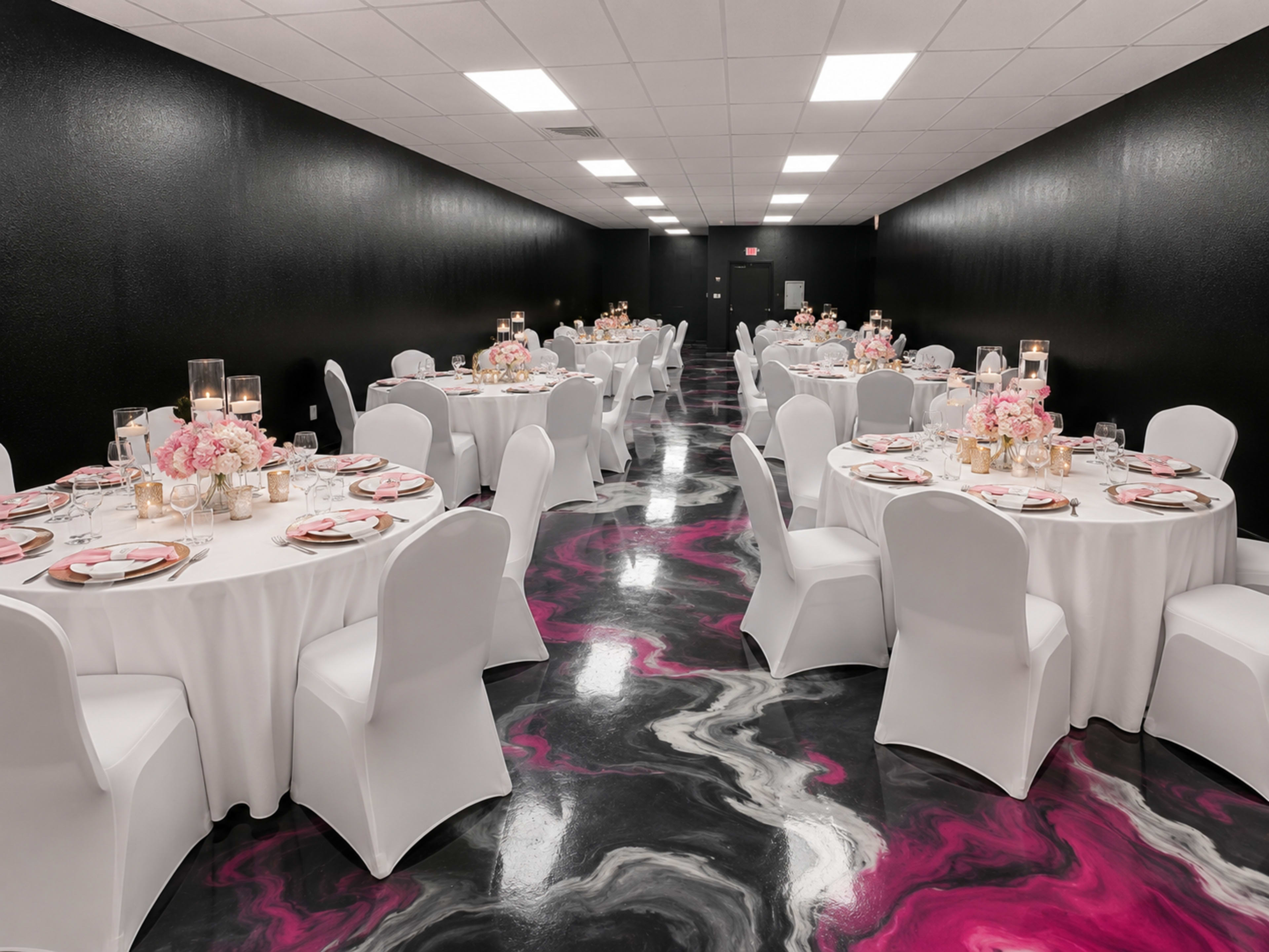 The image shows a stylish event space with neatly arranged round tables covered in white tablecloths, adorned with pink floral centerpieces and lit candles, all set against a black wall and a colorful marbled floor.