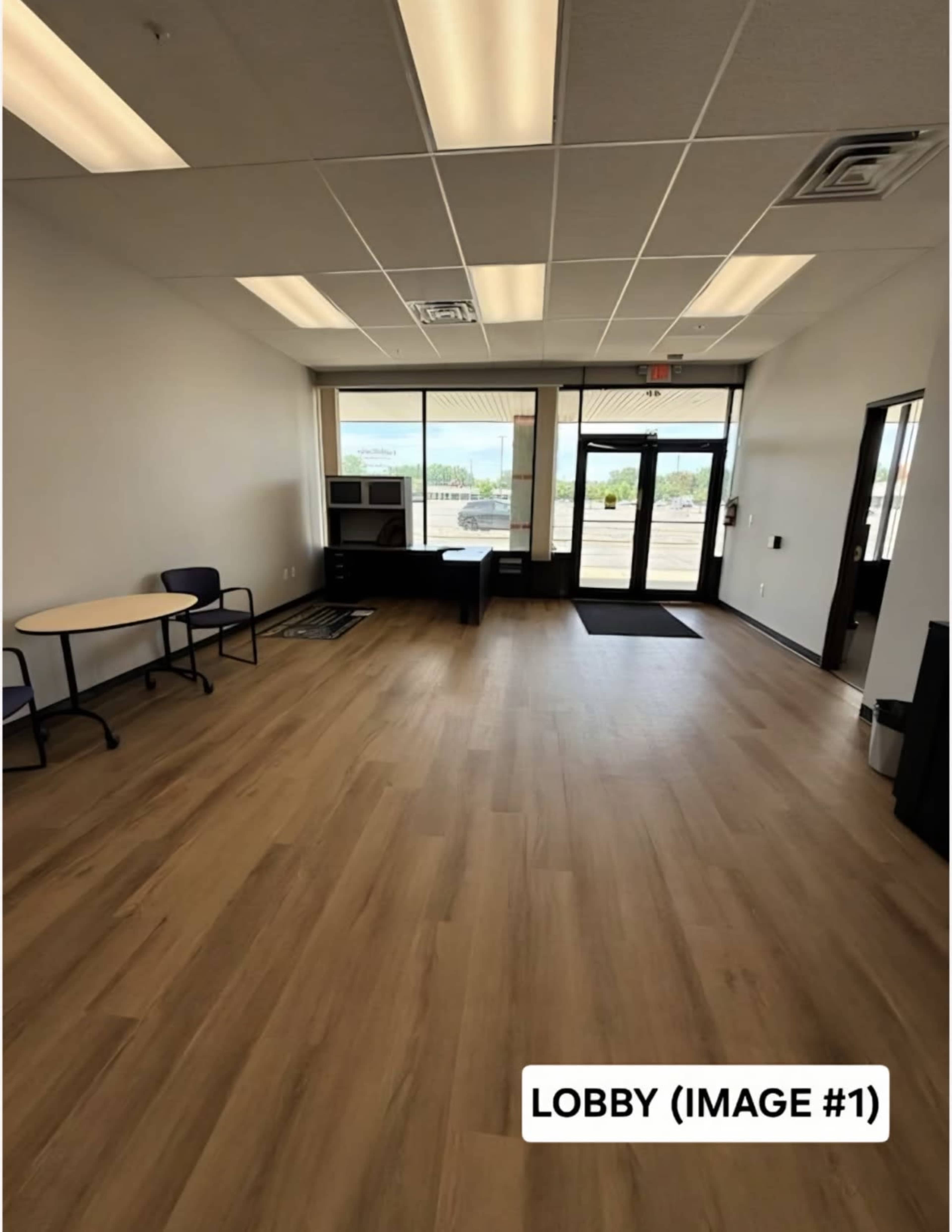 The image shows a spacious lobby area with wooden flooring, a small table and chairs, and a large window overlooking an outdoor view.