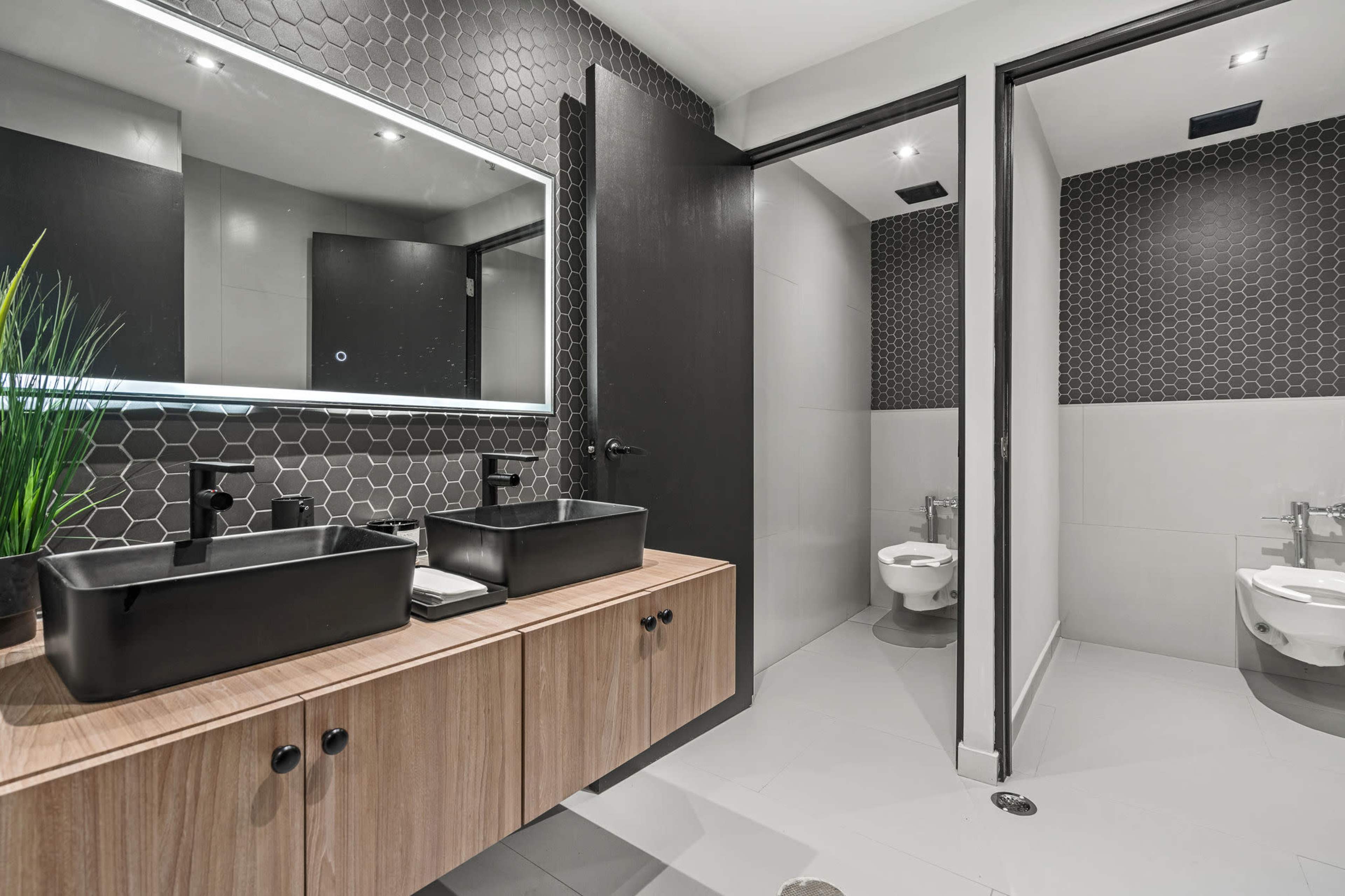 The image shows a modern bathroom with a dual sink vanity, black basin sinks, and two separate toilet stalls situated behind a wall.
