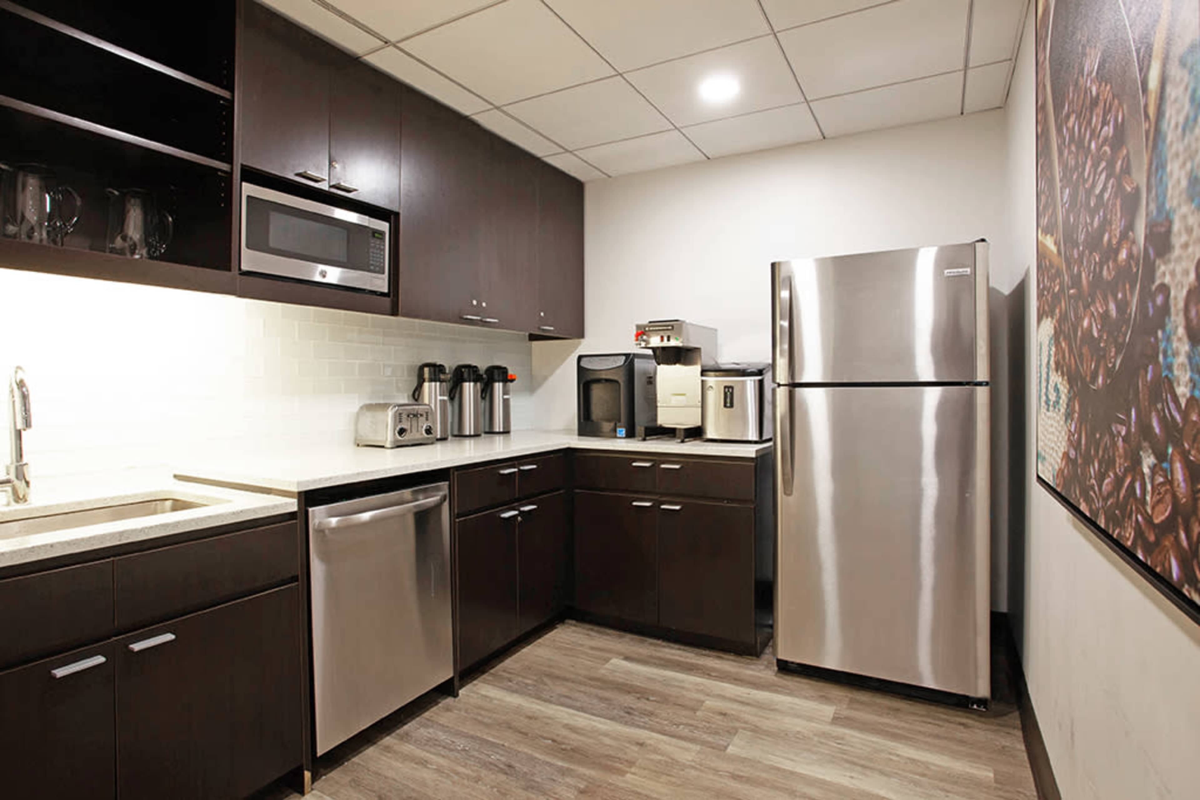 A modern kitchen with dark cabinetry, a stainless steel refrigerator, a microwave, and various appliances neatly arranged on countertops.