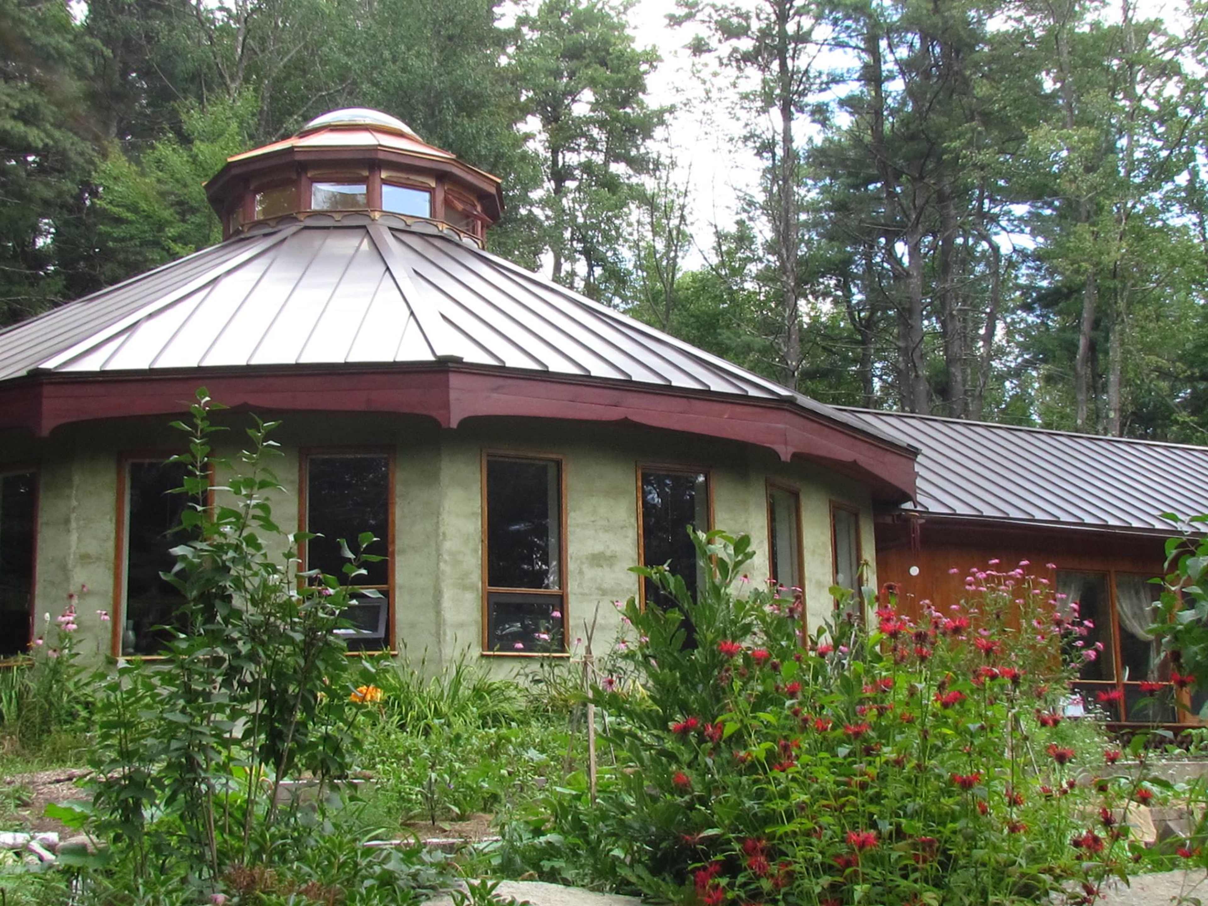 A round, multi-windowed building with a metal roof is surrounded by vibrant gardens and lush trees.