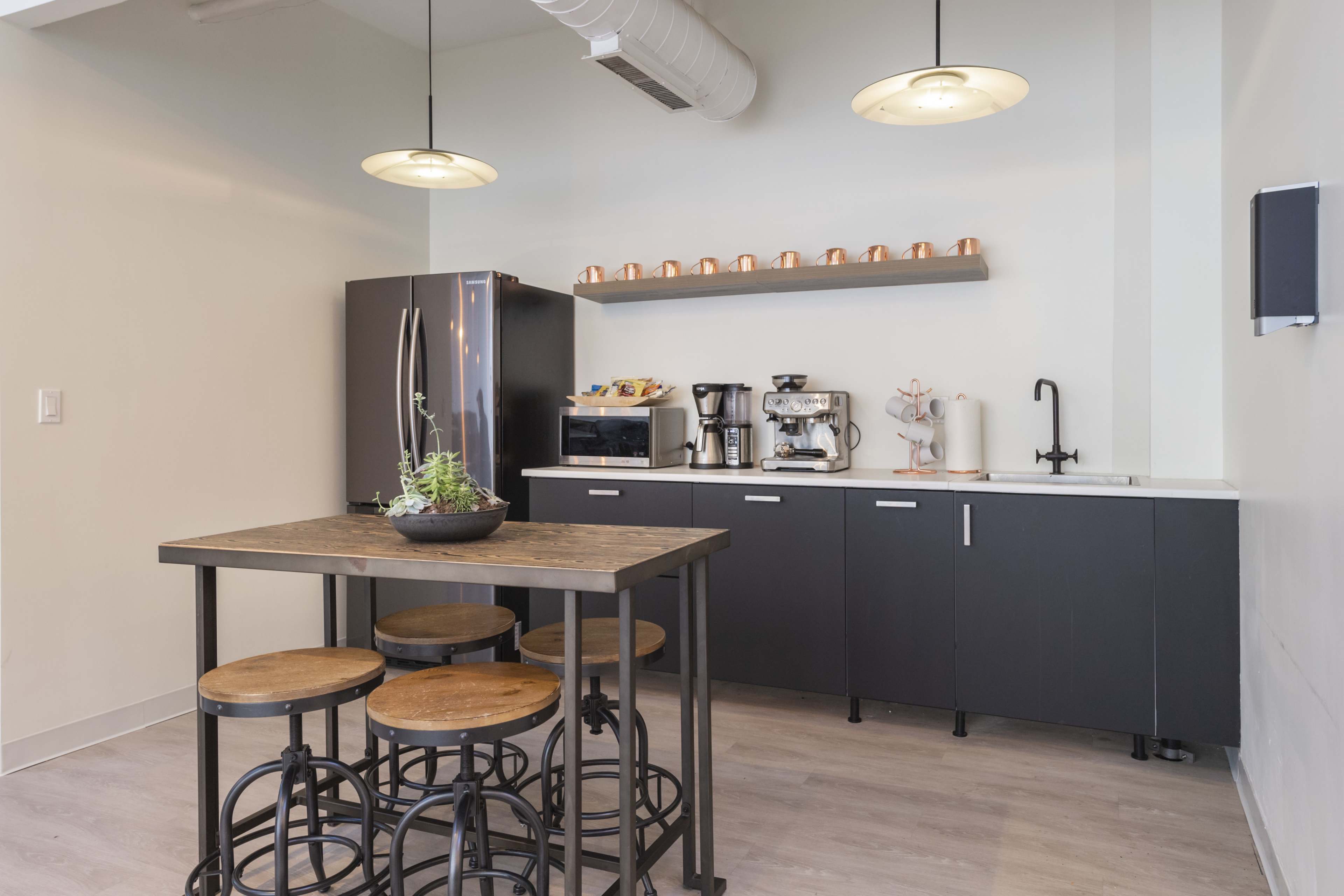 A modern kitchen area with a black refrigerator, coffee machines, and a wooden table surrounded by stools.