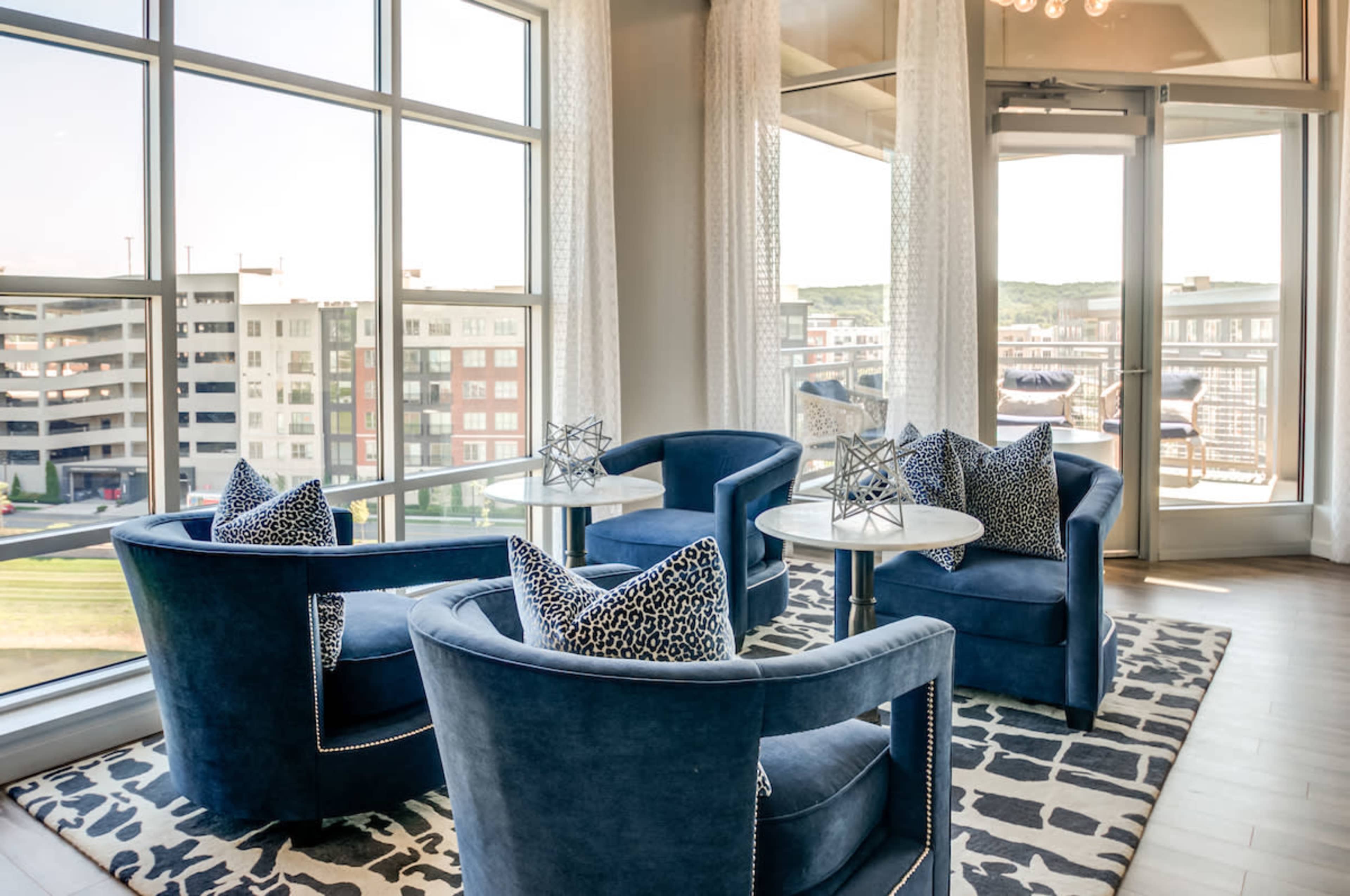 A stylish seating area features two blue armchairs with decorative pillows and glass-topped tables, surrounded by large windows overlooking a parking structure and nearby buildings.