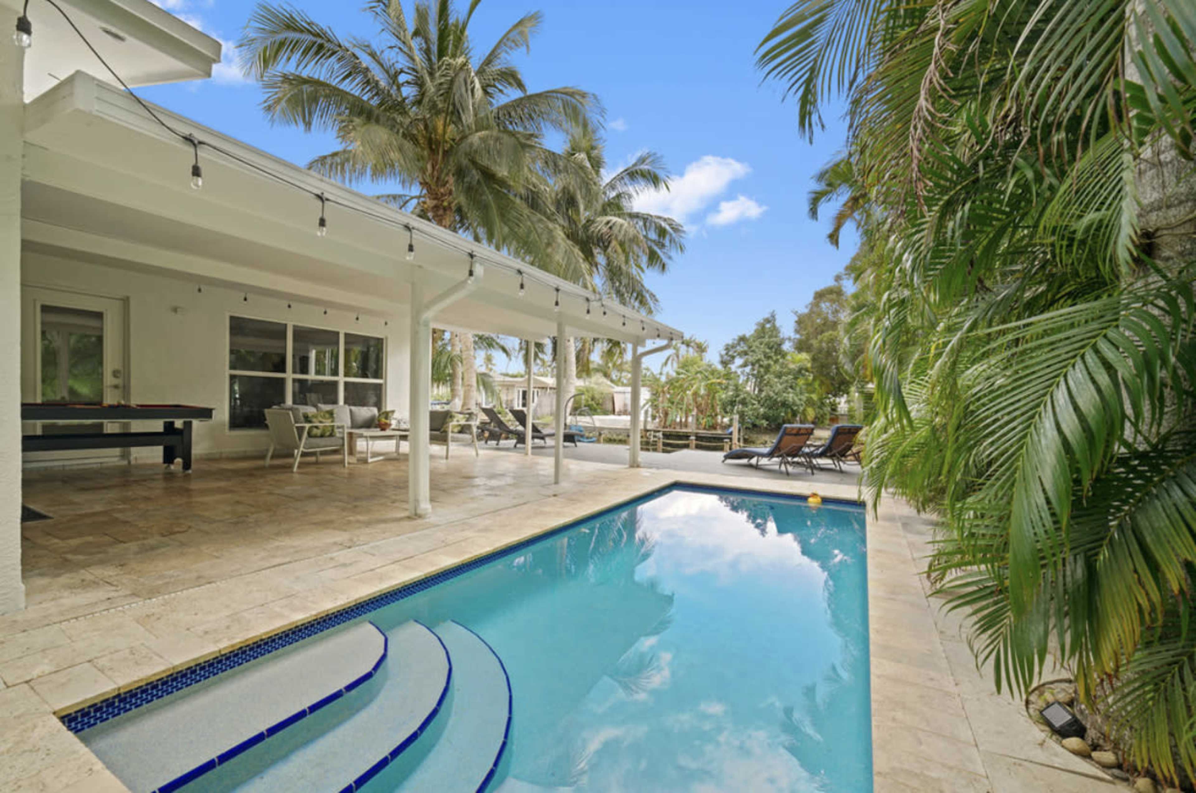 A swimming pool surrounded by a stone patio, with palm trees and lounge chairs in a sunny outdoor setting.