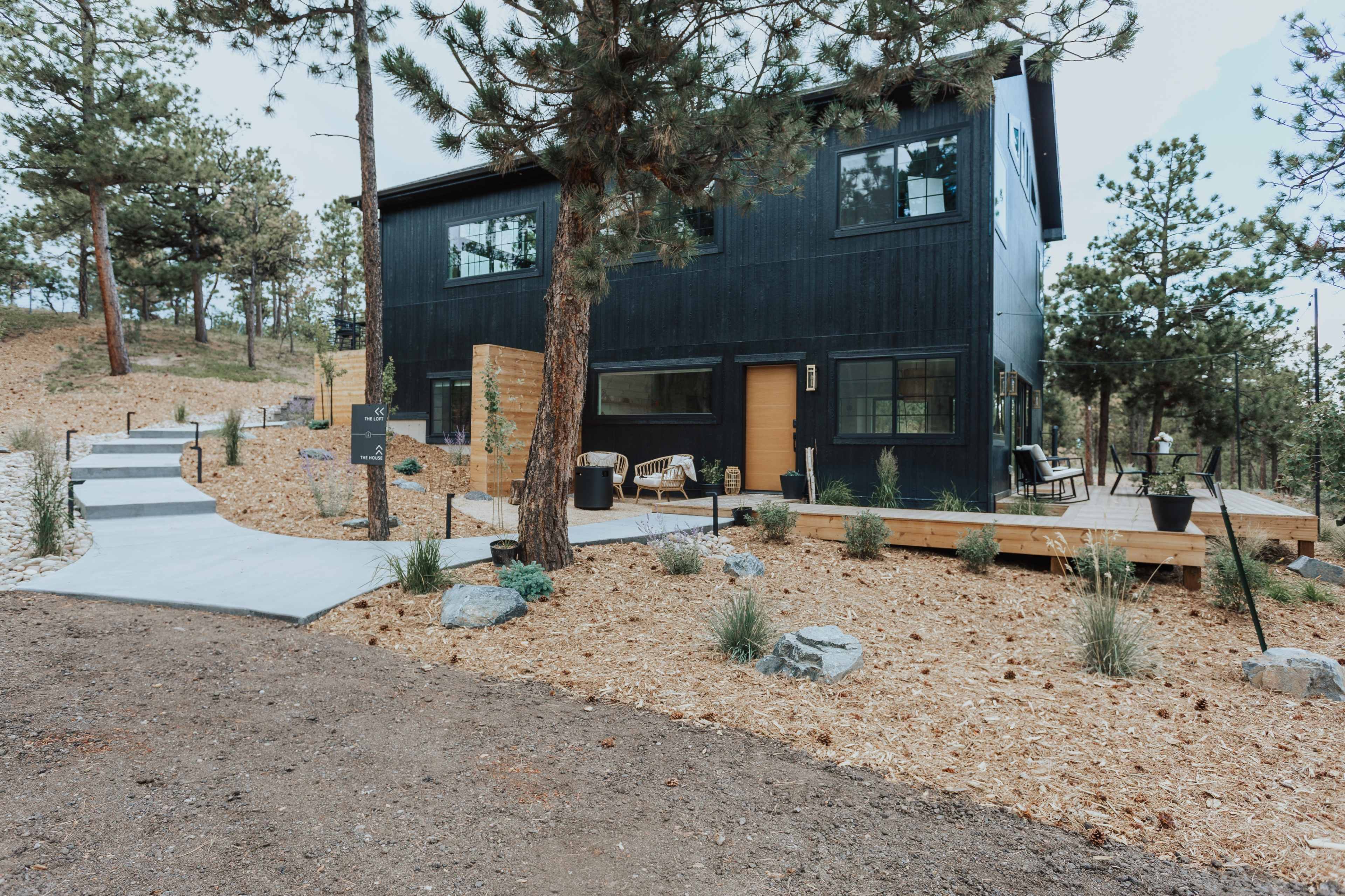 A modern black house with large windows stands surrounded by pine trees and landscaping featuring gravel paths and shrubs.