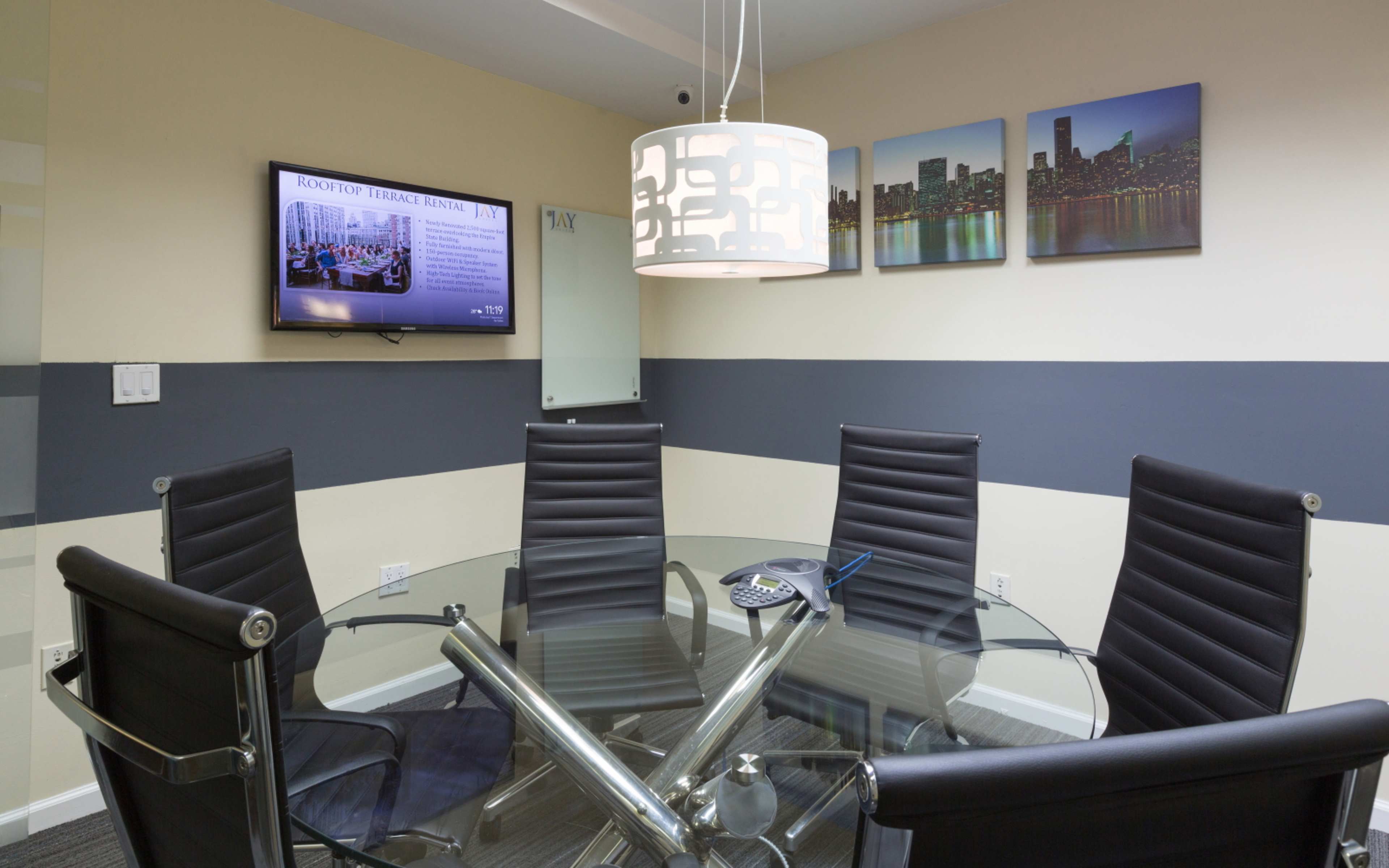 A modern conference room with a glass table, black leather chairs, a television mounted on the wall, and cityscape artwork.