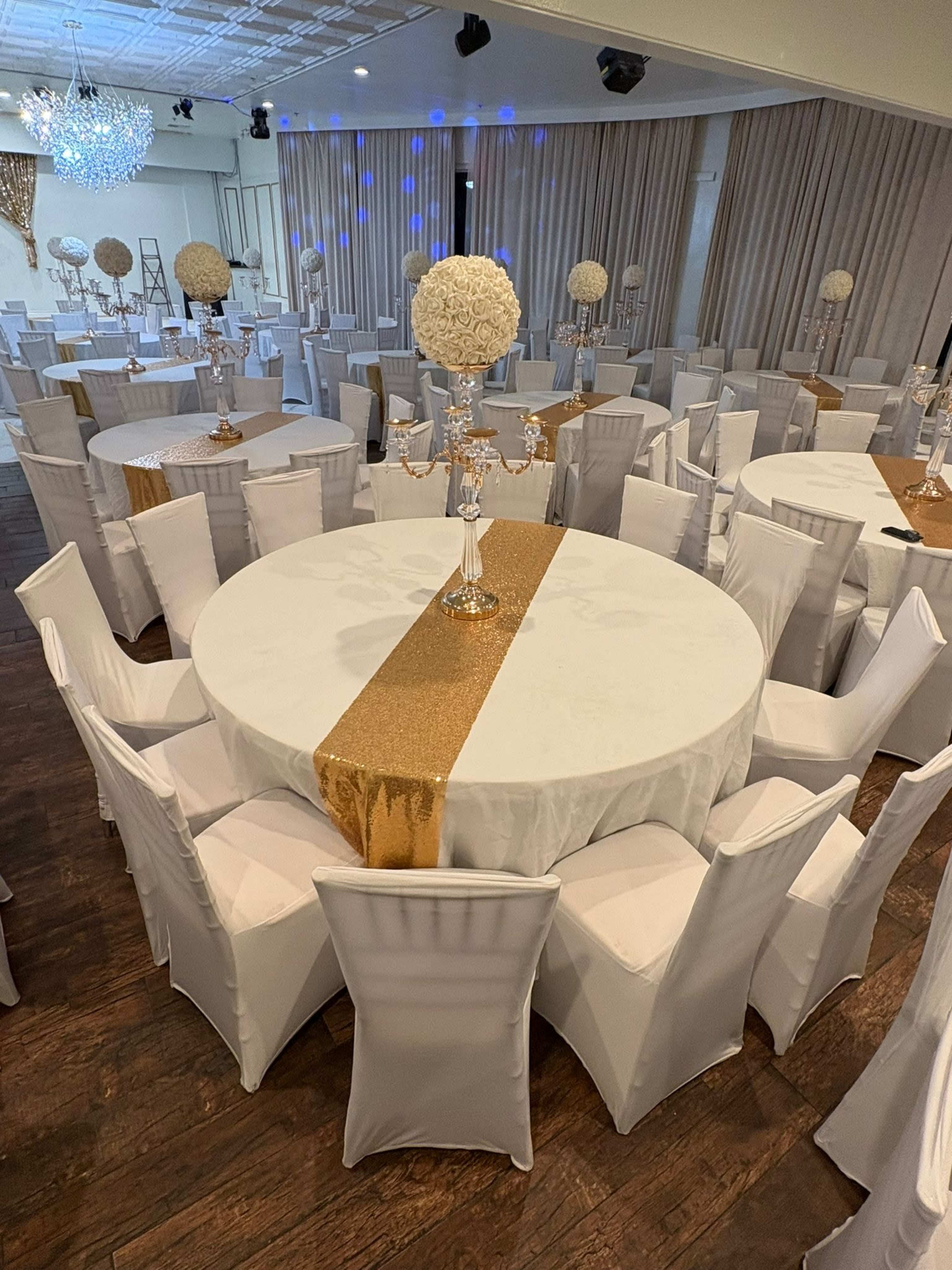 The image shows a decorated banquet hall with round tables covered in white cloth and gold accents, surrounded by white chairs, with floral centerpieces on each table.