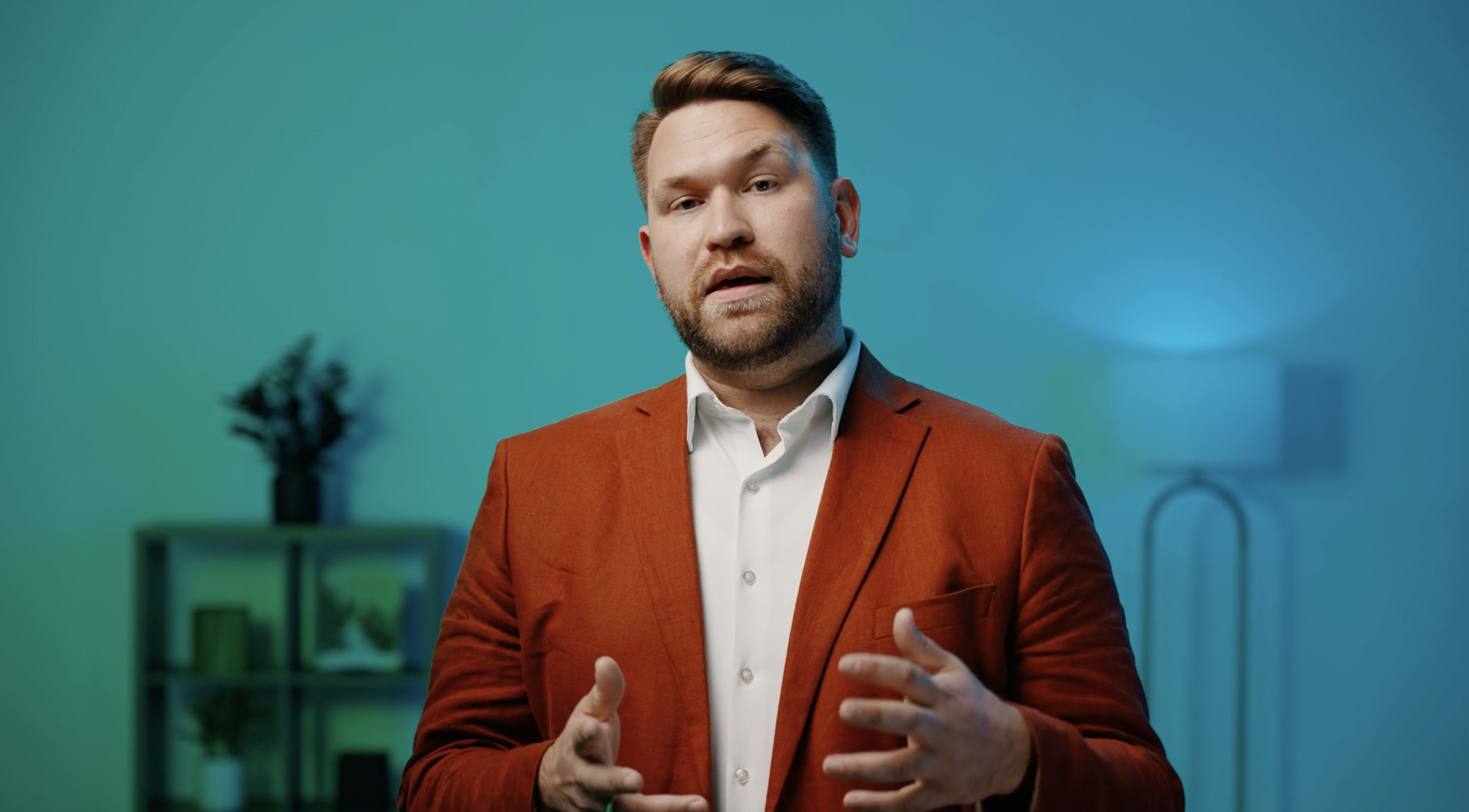A man in a red blazer gestures while speaking against a blue-green background.