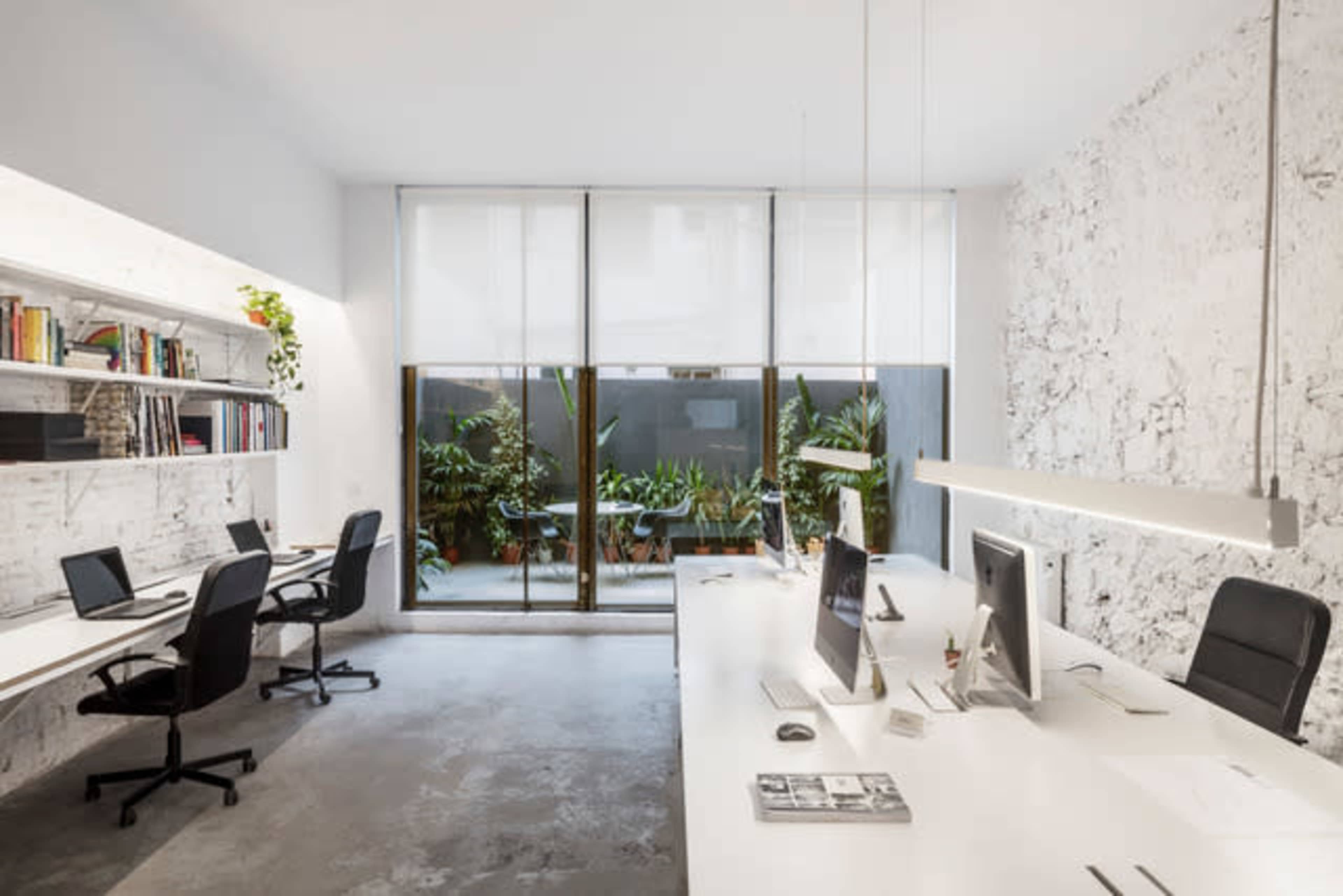 A modern office space featuring two workstations with computers, shelves filled with books, and large windows opening to a green outdoor area.