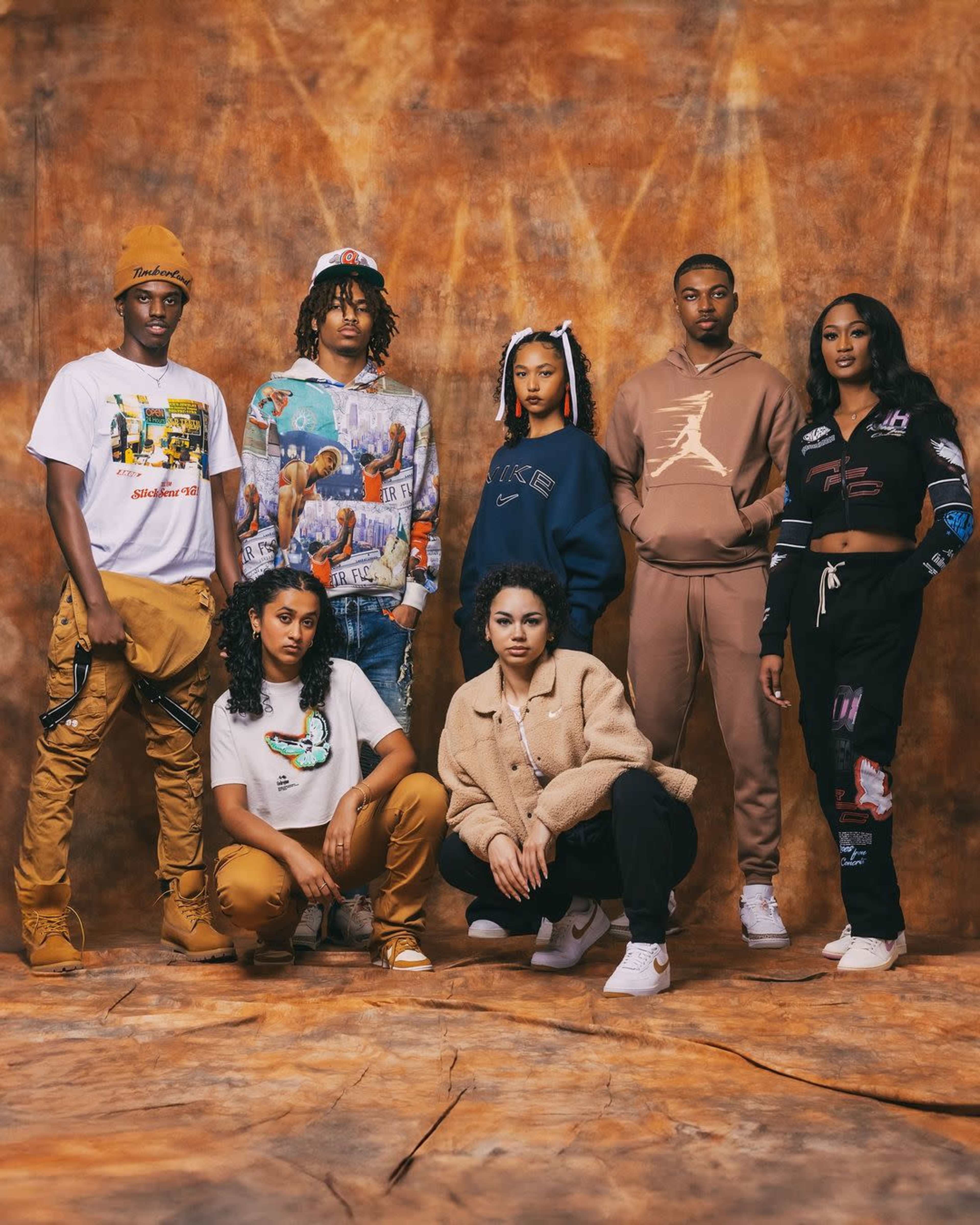 A group of seven young people poses together in stylish casual clothing against a textured brown backdrop.