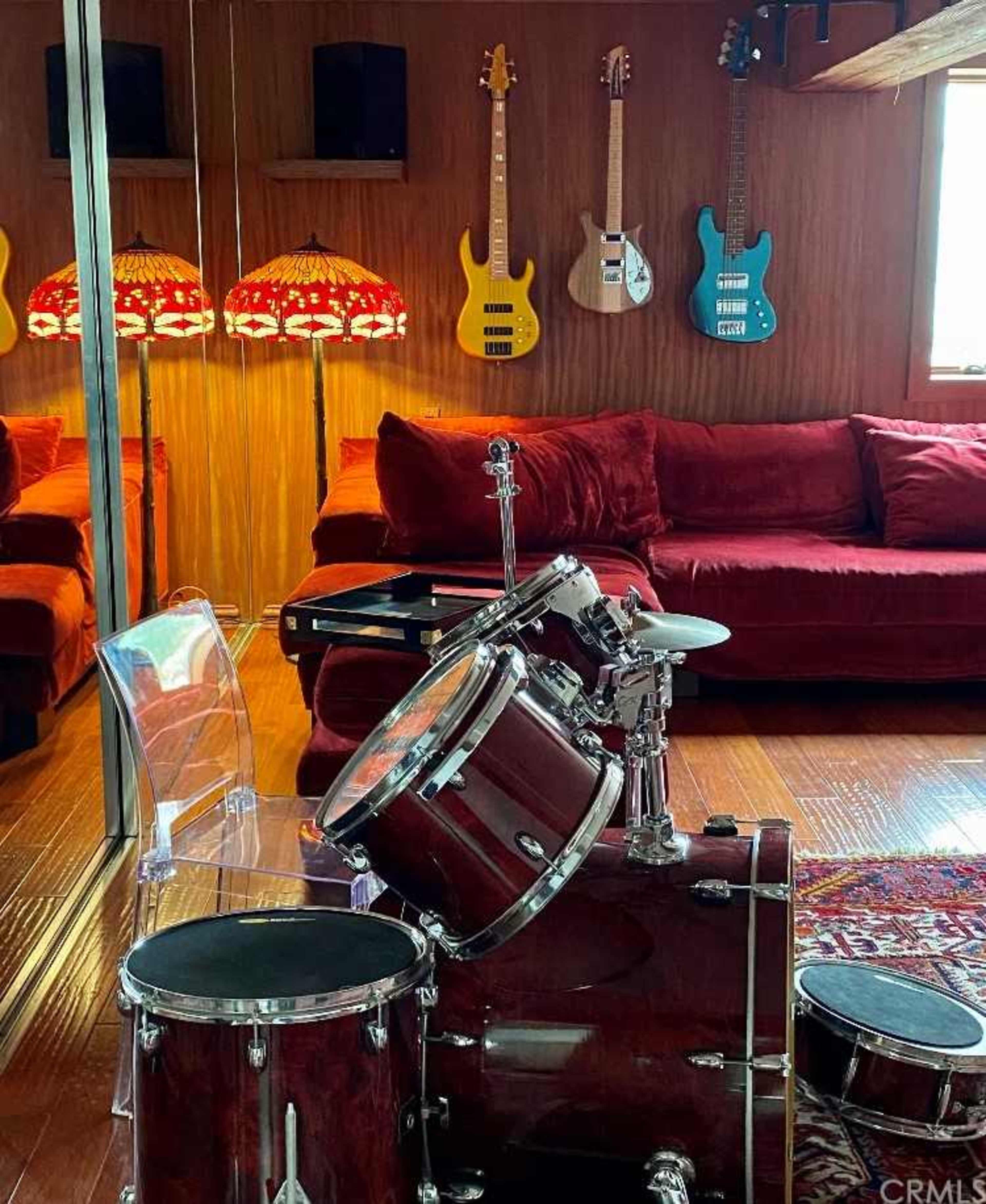 A room featuring a drum set in the foreground, a red velvet couch, and illuminated lamps, with electric guitars hanging on the wall in the background.