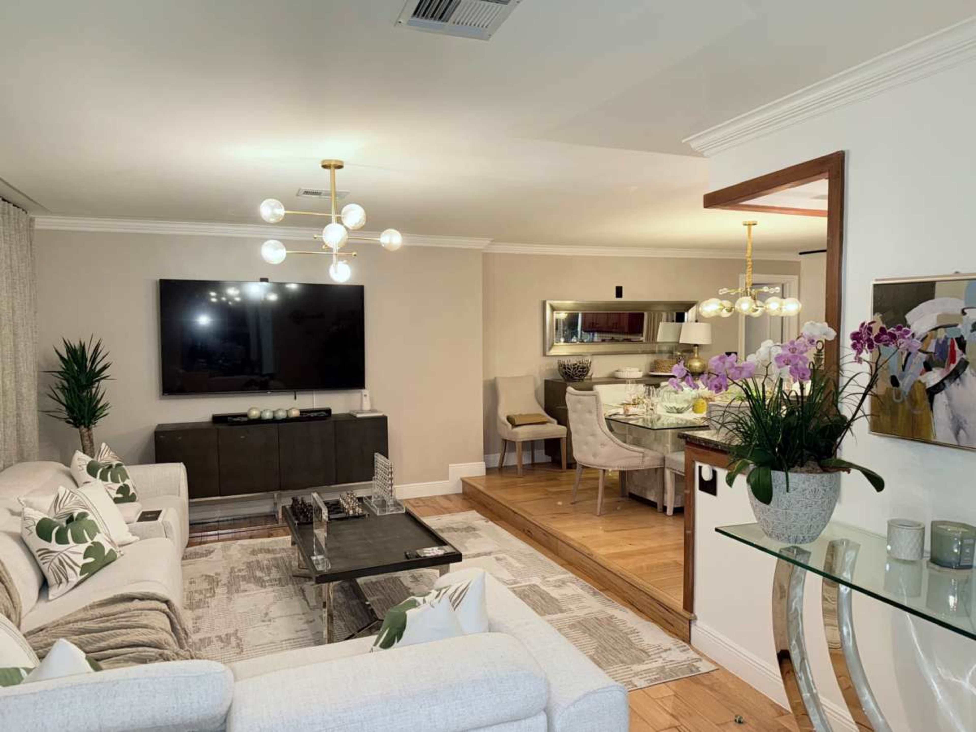 The image shows a modern living room with a television mounted on the wall, a seating area, and a dining space in the background.